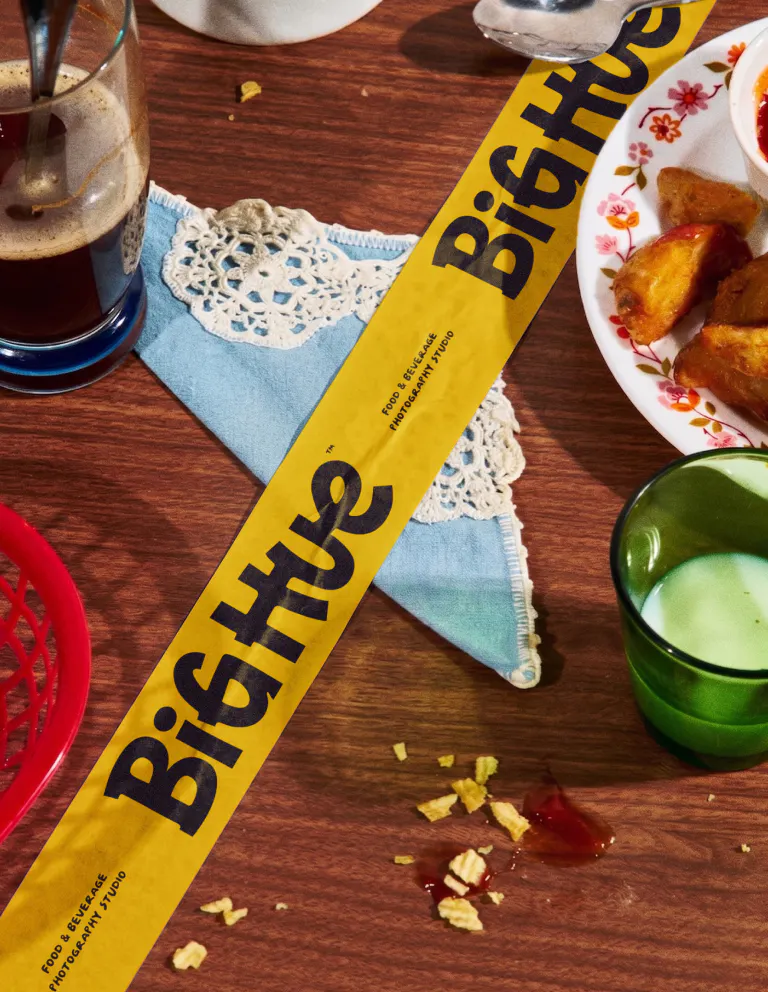 A yellow branded sash printed with the Big Hue wordmark and "Food & Beverage Photography Studio" laid diagonally across a wooden diner table. Surrounded by iced coffee, fried snacks, a crocheted napkin, and scattered crumbs.