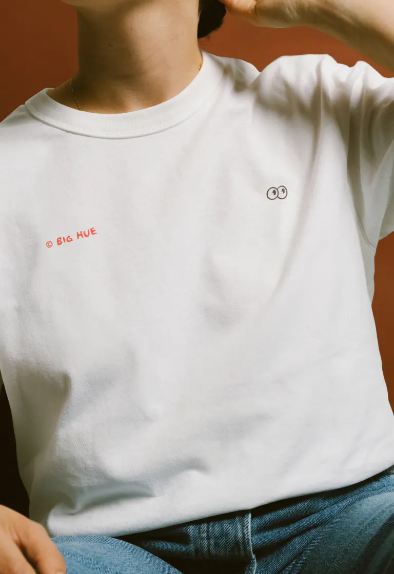 Close-up of a person wearing a white crewneck t-shirt. "© BIG HUE" printed in small red text on the left chest. The eyes mascot icon printed in dark ink on the right chest.