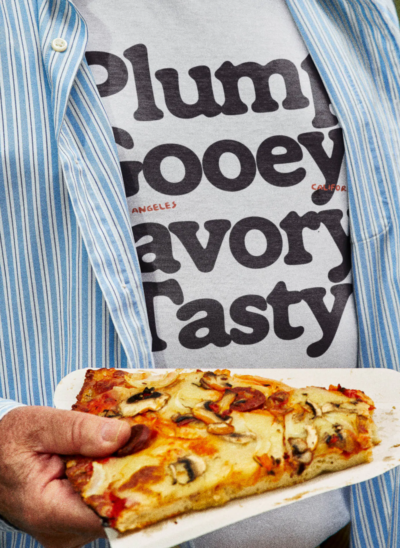 A person in a blue striped shirt holds a slice of square pizza. Underneath, a white t-shirt is visible printed with the stacked words "Plump / Gooey / Savory / Tasty" in the Ramboia Bold typeface. Small red text reads "Los Angeles, California."