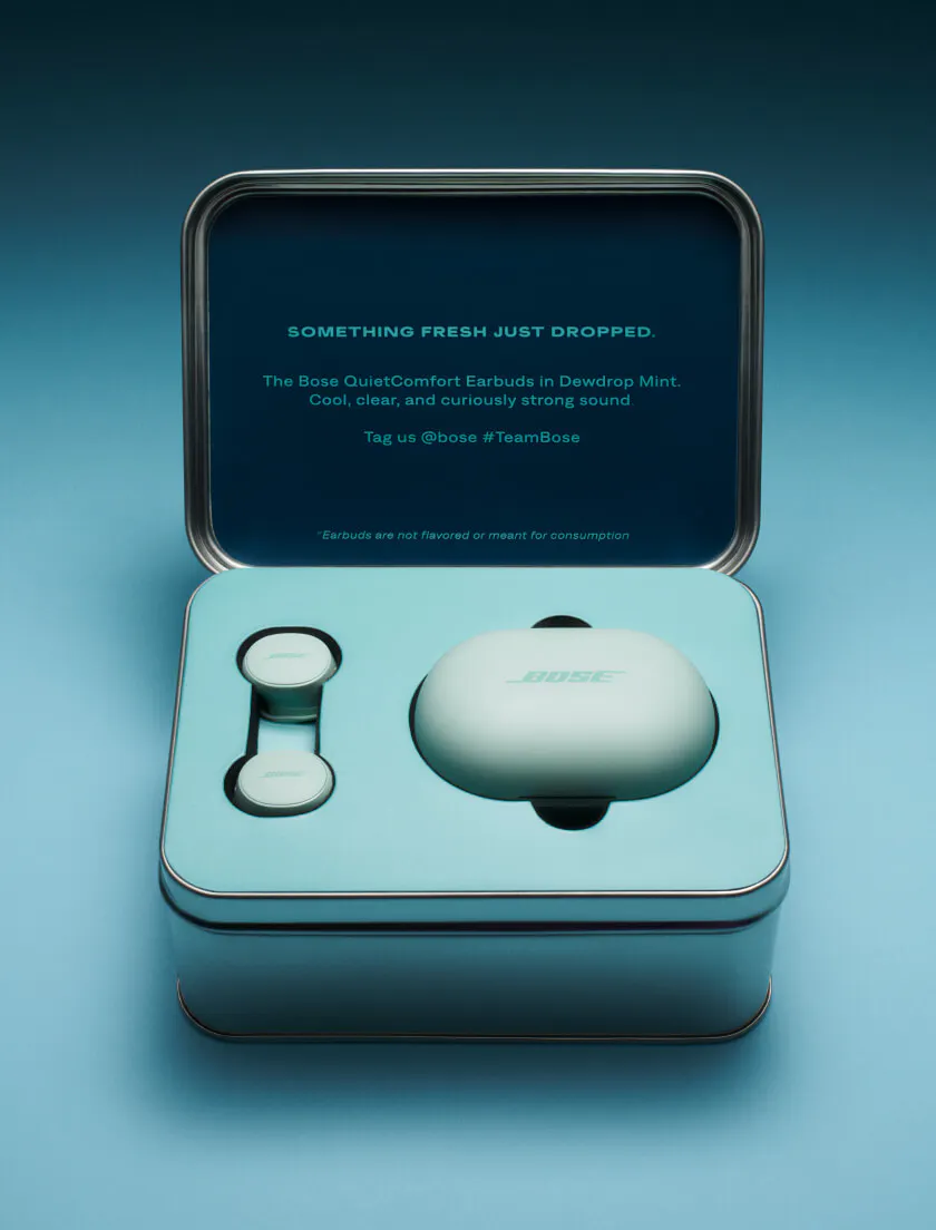 The Bose Dewdrop Mints seeding tin open on a teal background. The dark navy lid reads "Something Fresh Just Dropped — The Bose QuietComfort Earbuds in Dewdrop Mint. Cool, clear, and curiously strong sound." The earbuds and charging case sit nestled in mint-colored foam below.