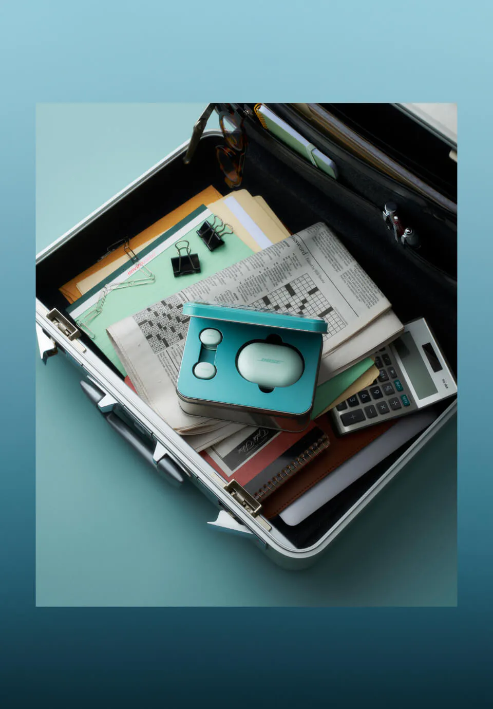 the open Bose QuietComfort Earbuds charging case resting on a cluttered briefcase filled with papers, a crossword, and a calculator.