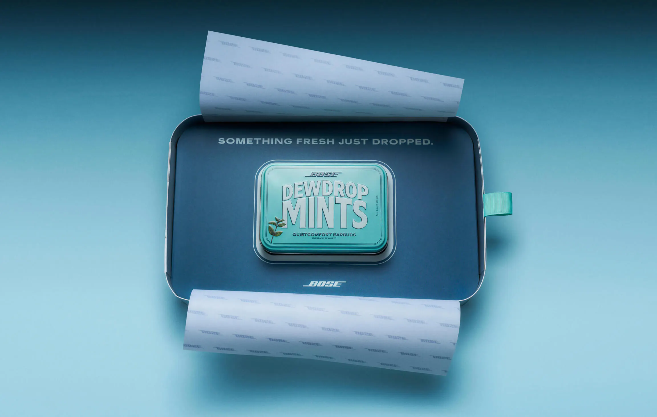 Top-down view of the Bose Dewdrop Mints seeding box opened flat, revealing the mint tin at center on a navy interior printed with "Something Fresh Just Dropped." Branded tissue paper curls out from both ends.