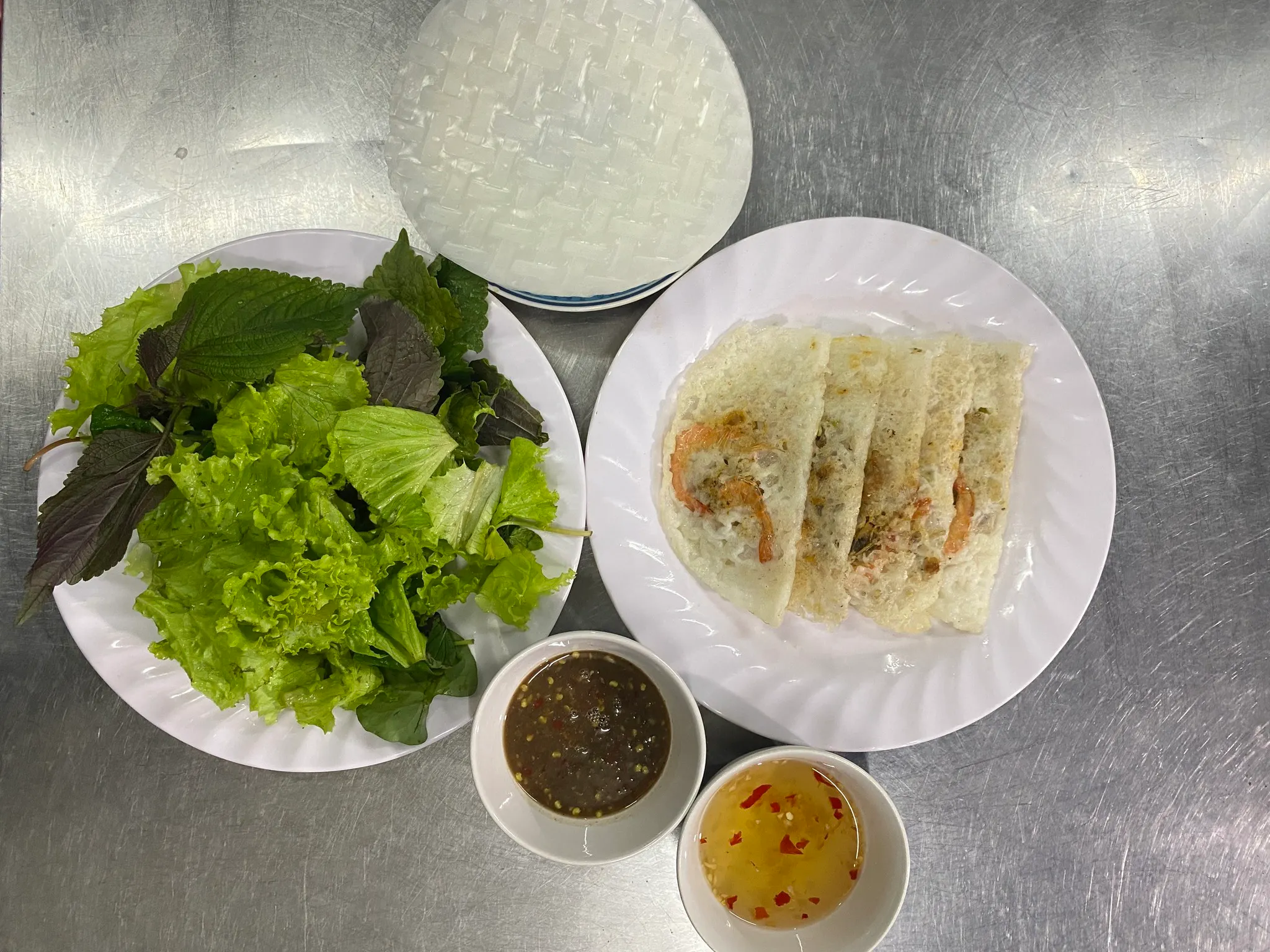 Hình ảnh mẹt bánh xèo vỏ xếp chồng lên nhau bên cạnh rổ rau sống xanh mướt và chén mắm đục giã tỏi ớt