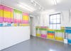 Installation view of 'Half' by henry Coleman, brightly colored posters overlap to horizontally  fill  half the available wall space of a small gallery space with other artworks mounted over the surface