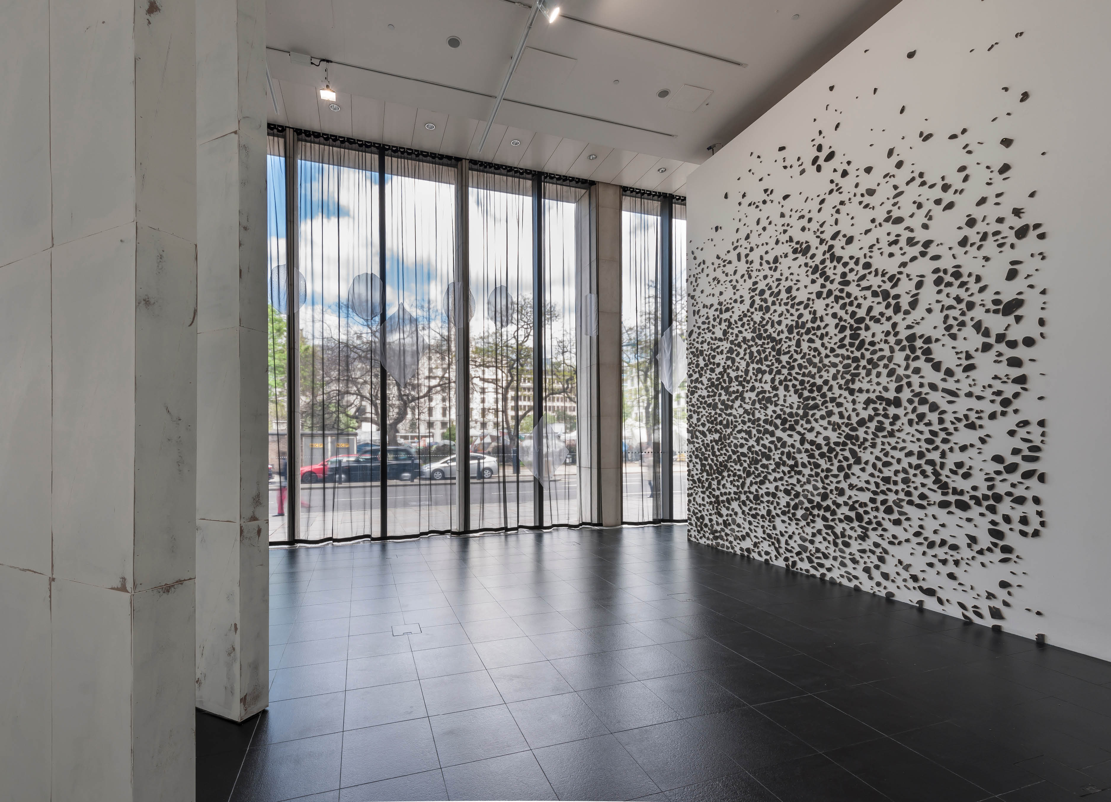 'About now' exhibition by Henry Coleman at Bloomberg SPACE, a large pattern of painted tiles are mounted across a wall in the pattern of a terrazo floor from the nearby Barbican Centre, A voile curtain with decorative elements runs across the windows 