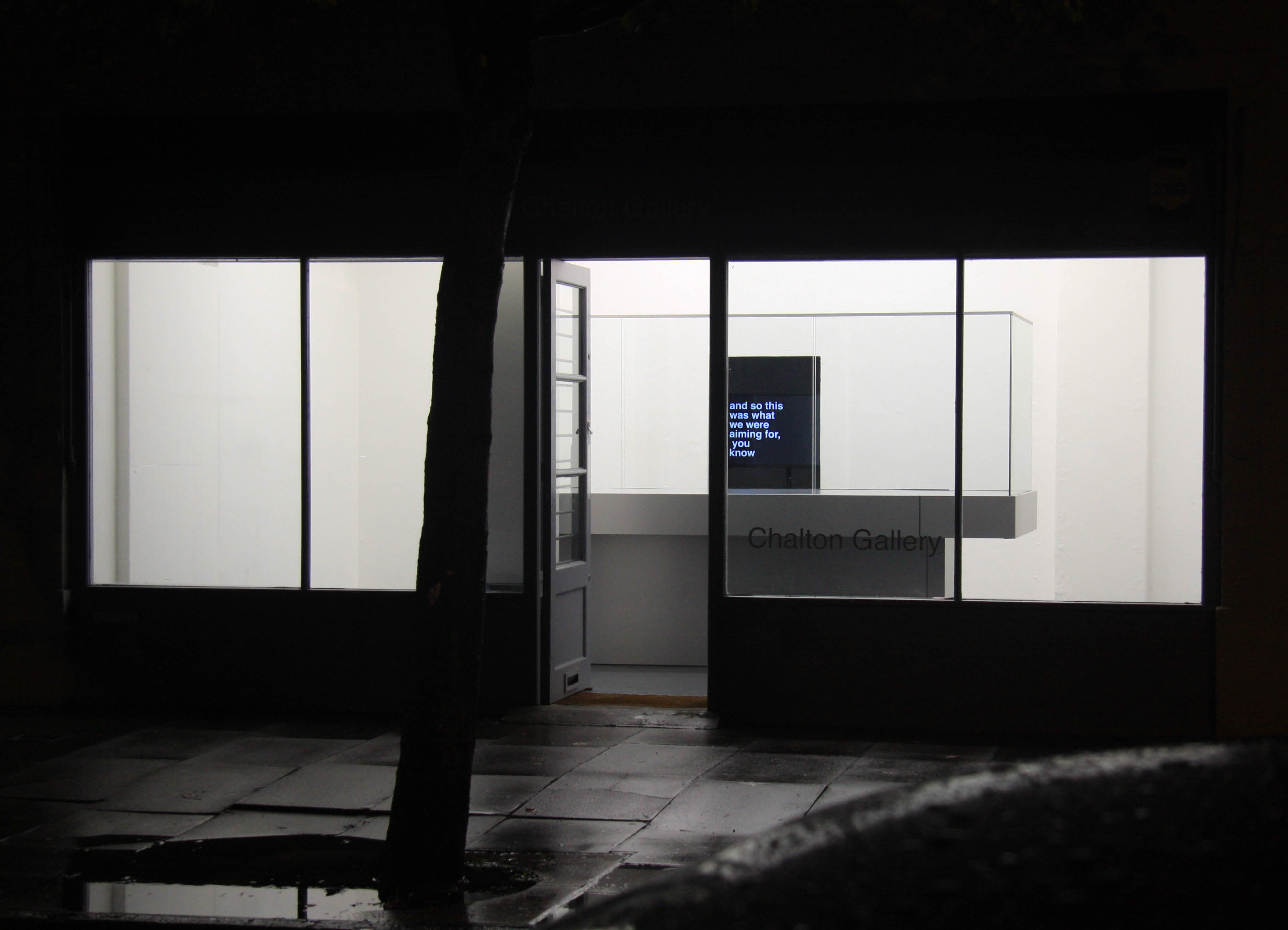 'My Sky' exhibition  by Artist Henry Coleman at Chalton Street gallery, lit  at night with a Sculptural installation of a glazed balcony structure with a film showing behind in front of an open doorway