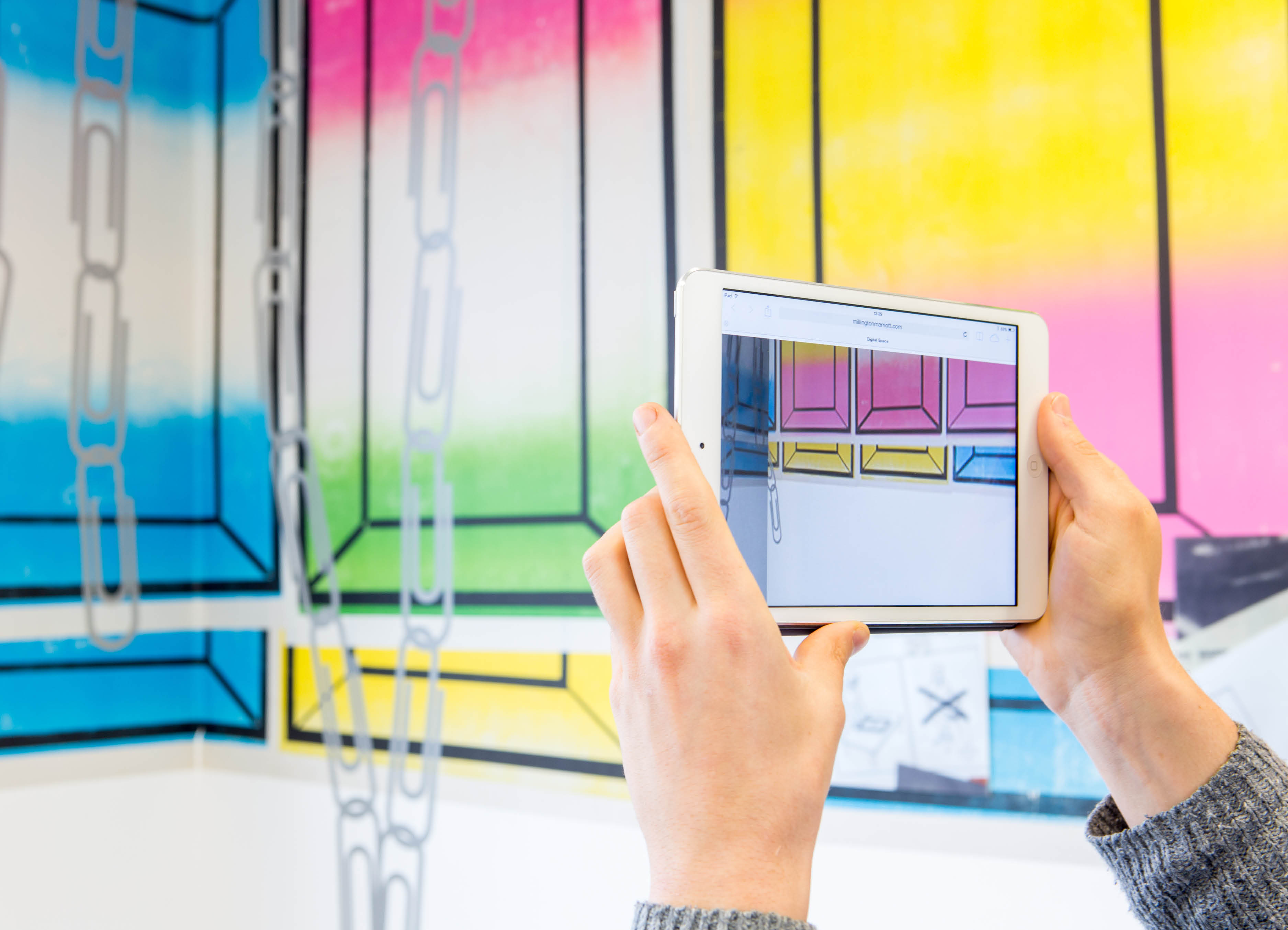 Augmented reality view of the artwork Half by henry Coleman, a pair of hands hold up an iPad to view an AR sculpture against a background of brightly colored posters