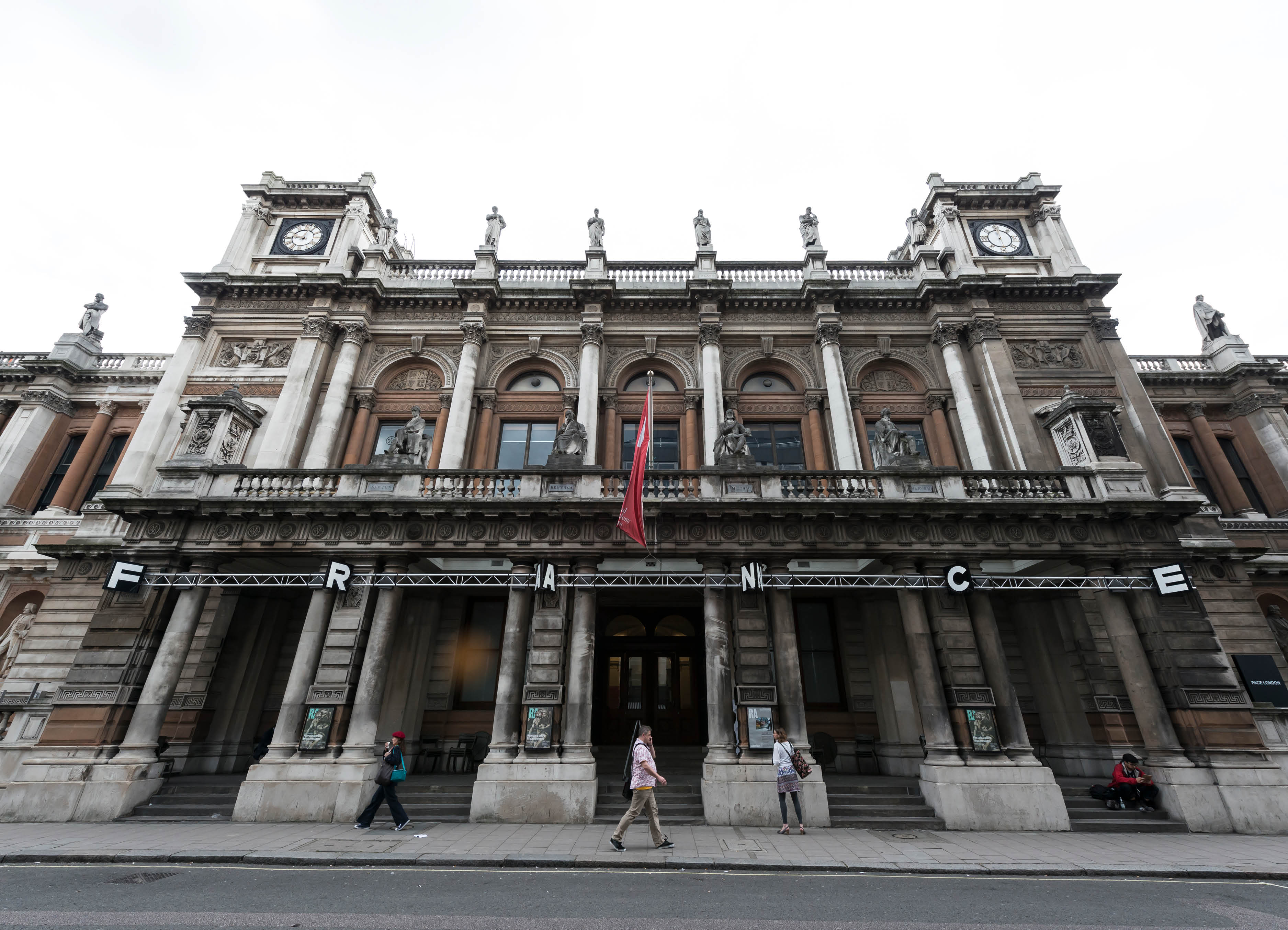 'Scrape' by artist Henry Coleman. Public art intervention of illuminated signage spelling out the word FRANCE attached to the facade of the RA building 6 Burlington Gardens, London at dusk with a man walking past on the phone