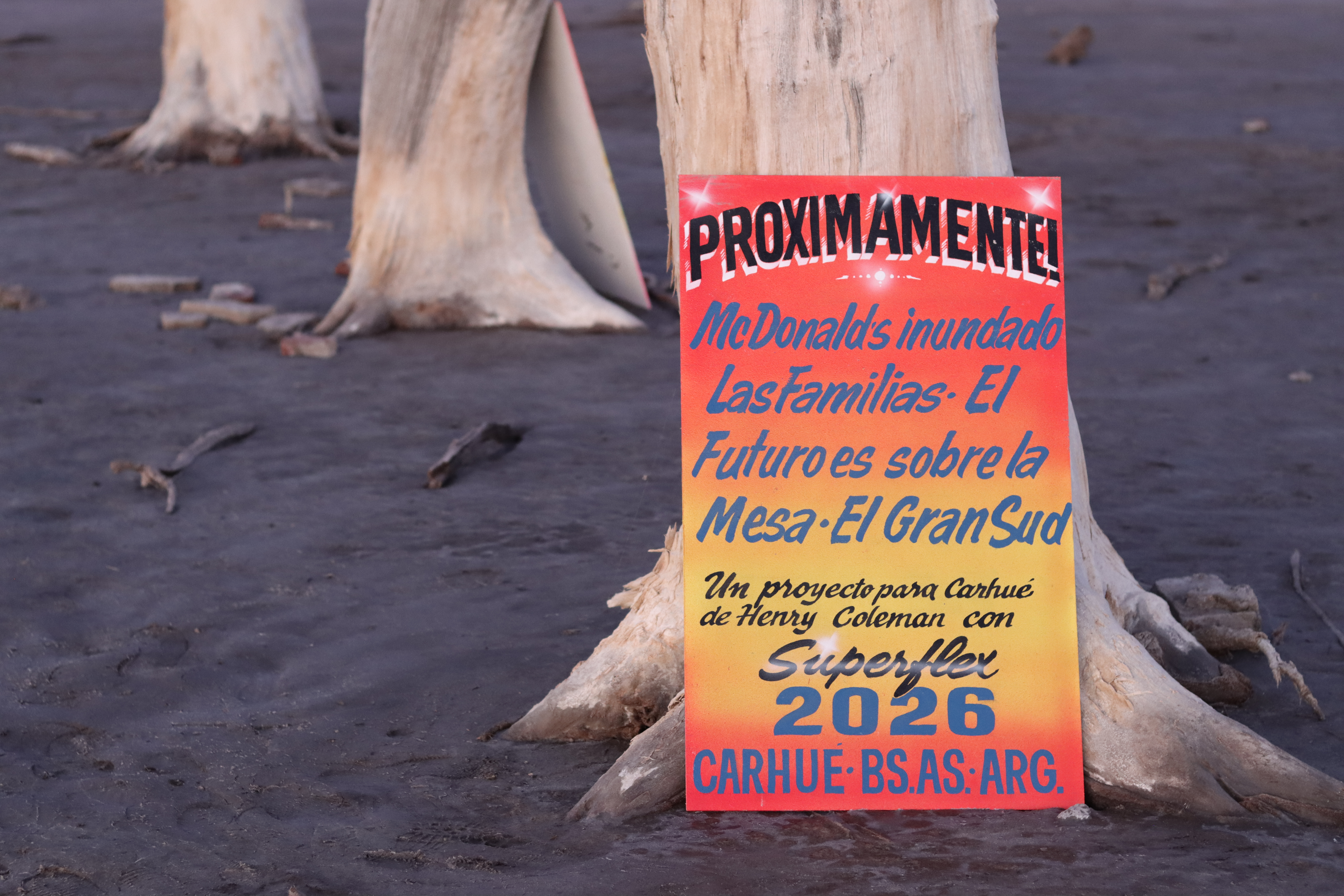 'PROXIMAMENTE!  by Henry Coleman with Colo Maldonado: Red and orange text board leaning on a tree by the side of Lago Epecuen.. 