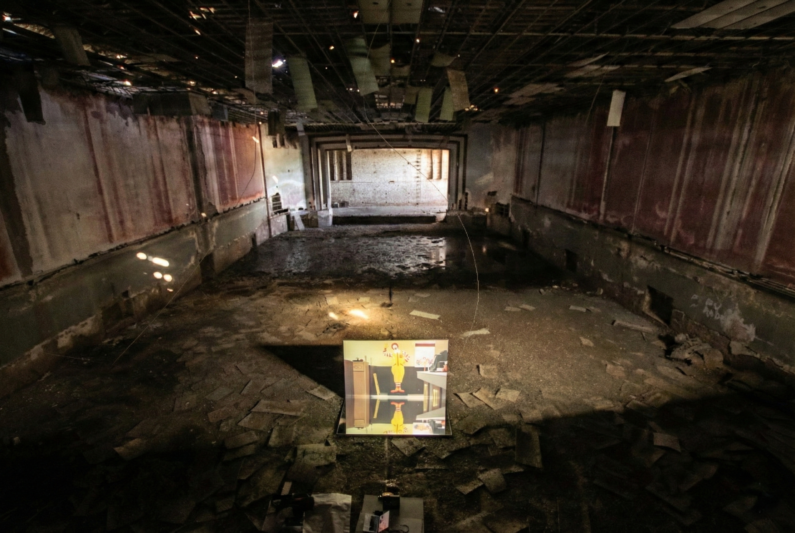 Flooded Doubled (2025) by Henry Coleman: Brightly lit video panel featuring Flooded McDonalds by Superflex with Ronald McDonald reflected on dark interior floor of Cinema Grand Sud.