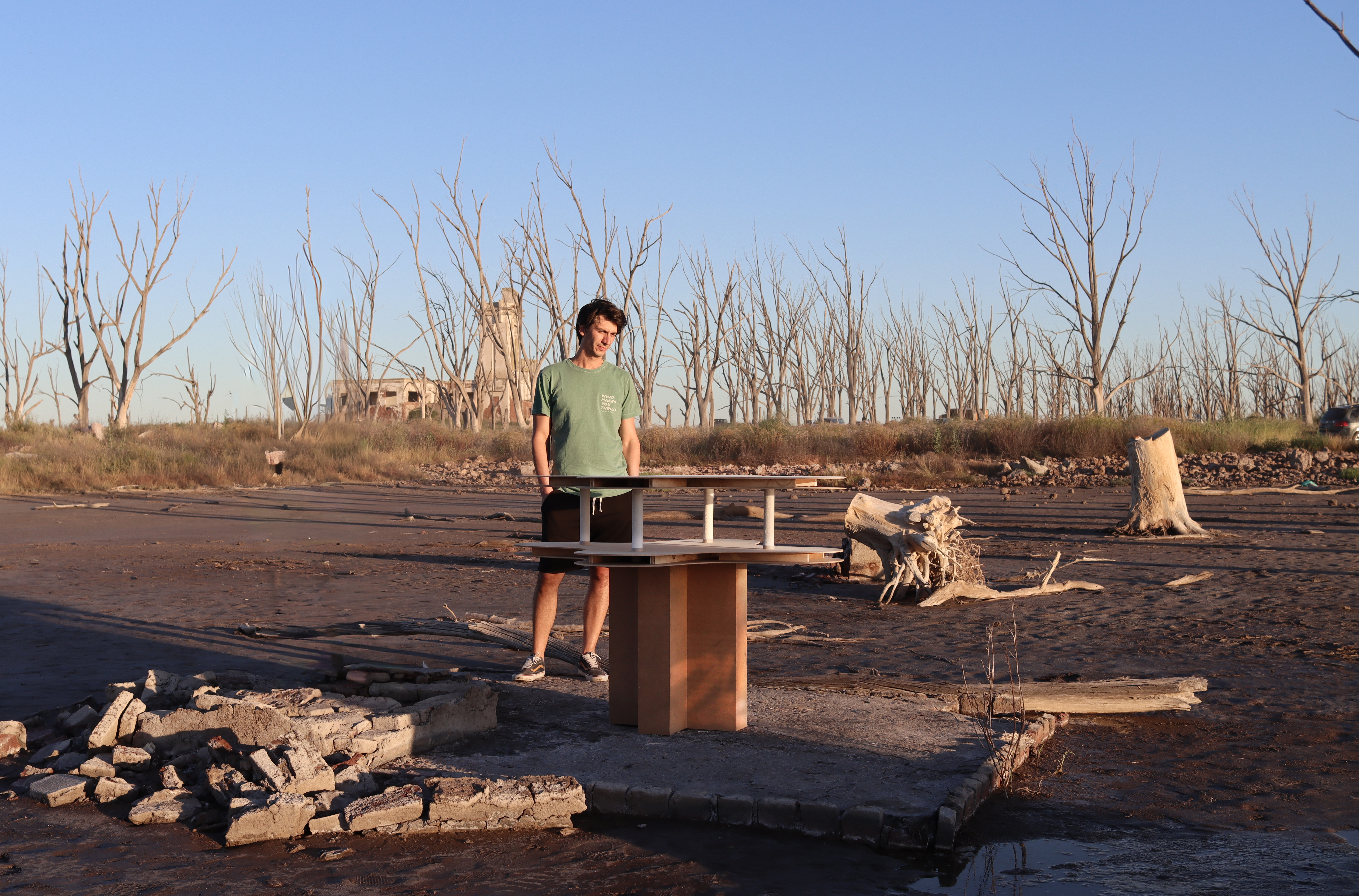 : El Futuro es sobre la Mesa (2025) by Henry Coleman: Man in green shirt looks down at multi-tiered wooden table form in the shape of the Ariston Cafe by Marcel Breur. Framed Against MAtadero by Salamone in  barren Epecuén landscape. 