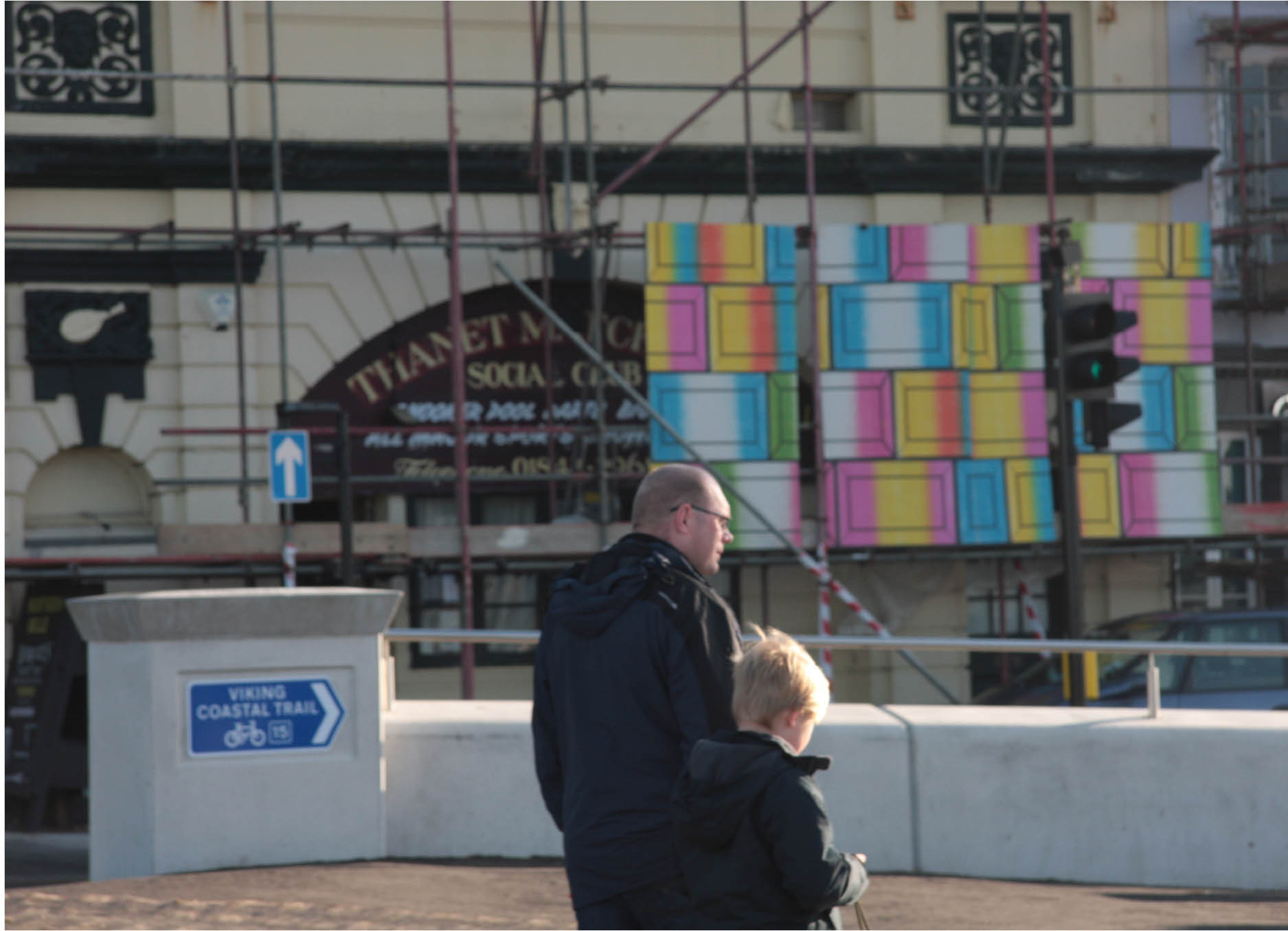 Public art installation by Henry Coleman as part of the World in a Box organized by Pete Fillingham in Margate, Kent, a hording of brightly colored posters is mounted on scaffolding in front of the Thanet Social club and a blue and white sign spells out Viking coastal route whilst a man and booty walk past in the foreground 