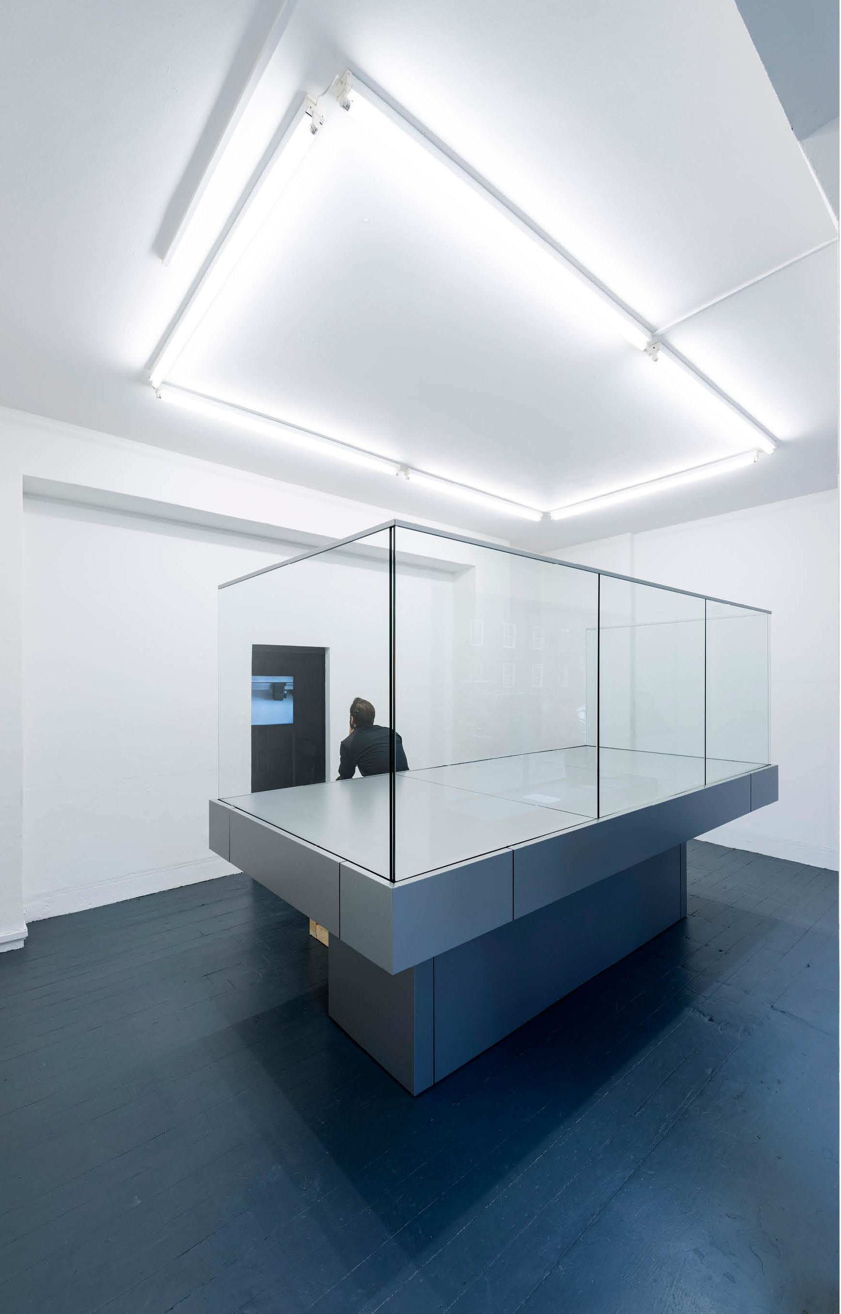 'My Sky' exhibition  by artist Henry Coleman at Chalton Street gallery, a man sits on the edge of a glazed balcony sculpture watching a film through a doorway under fluorescent lights