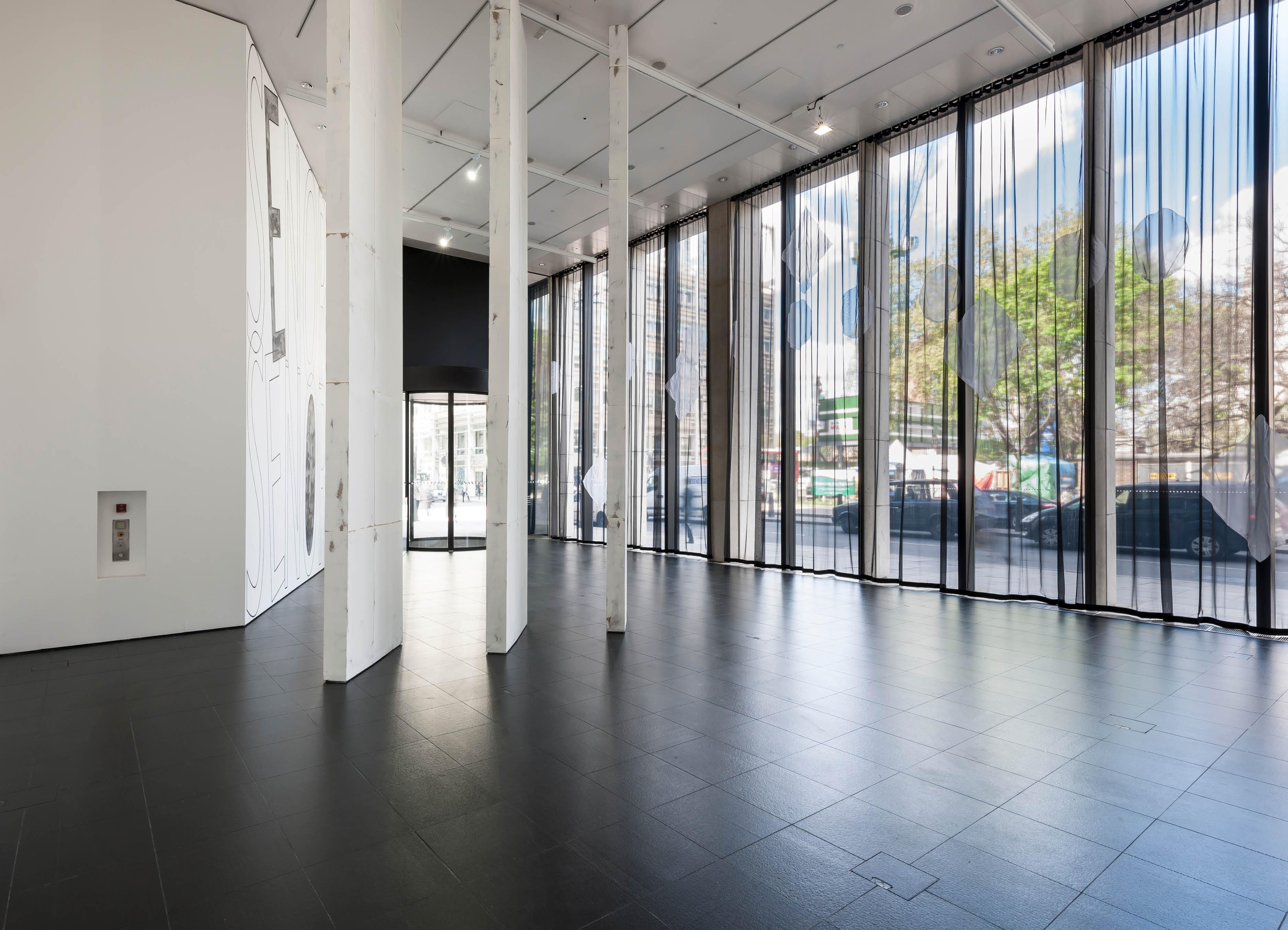 'looking through the 'About now' exhibition by Henry Coleman at Bloomberg SPACE, A voile curtain with decorative elements runs across the windows and three large mullion sculptures based on the form of Swiss cottage library divide the space 