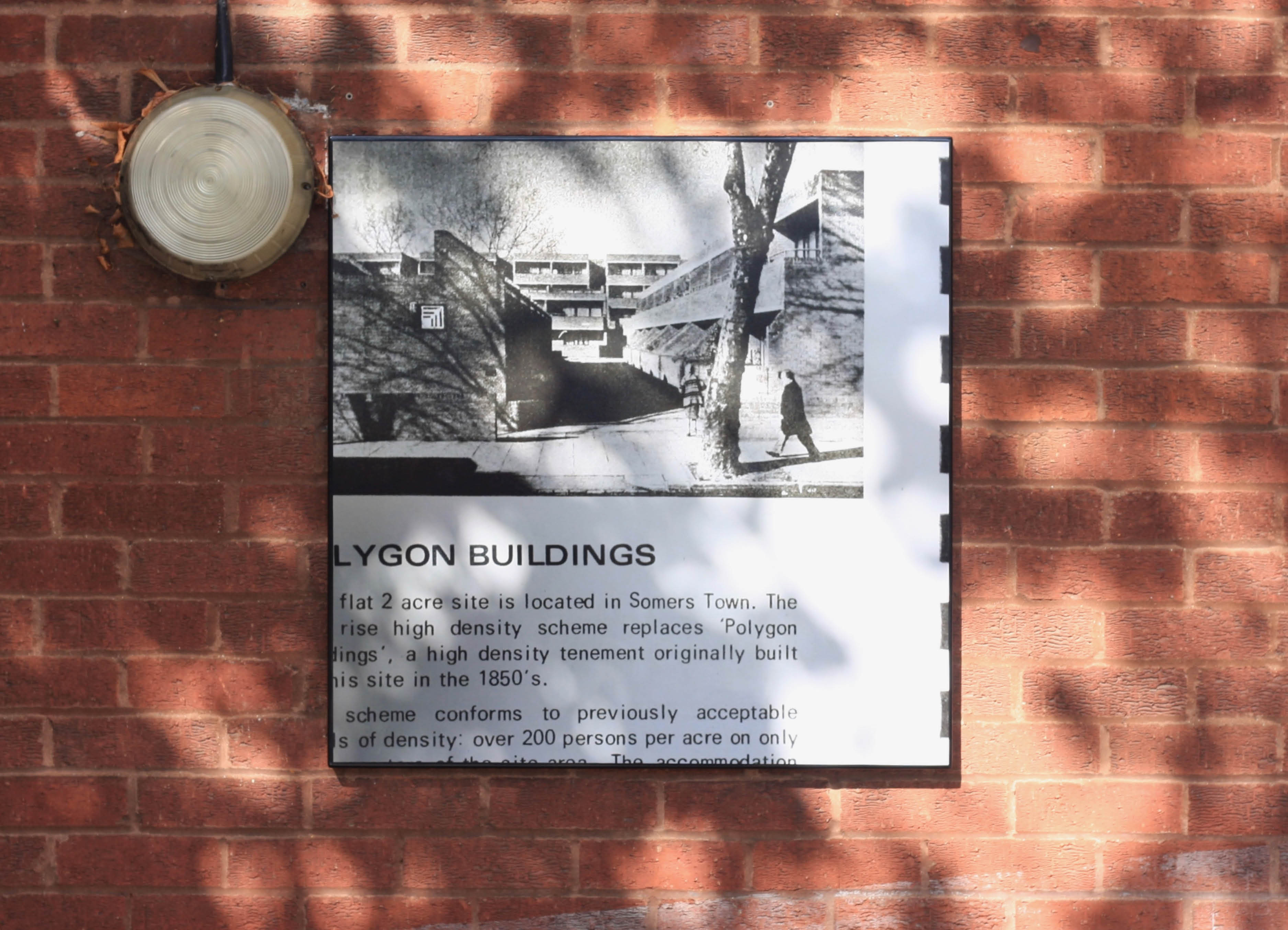 Polygon Buildings' by public artwork Henry Coleman, A detail of  a black and white picture of a man walking past the modernist polygon estate in Somers Town by Architect Tabori is mounted on a brick wall, text partially spells out Polygon Buildings 