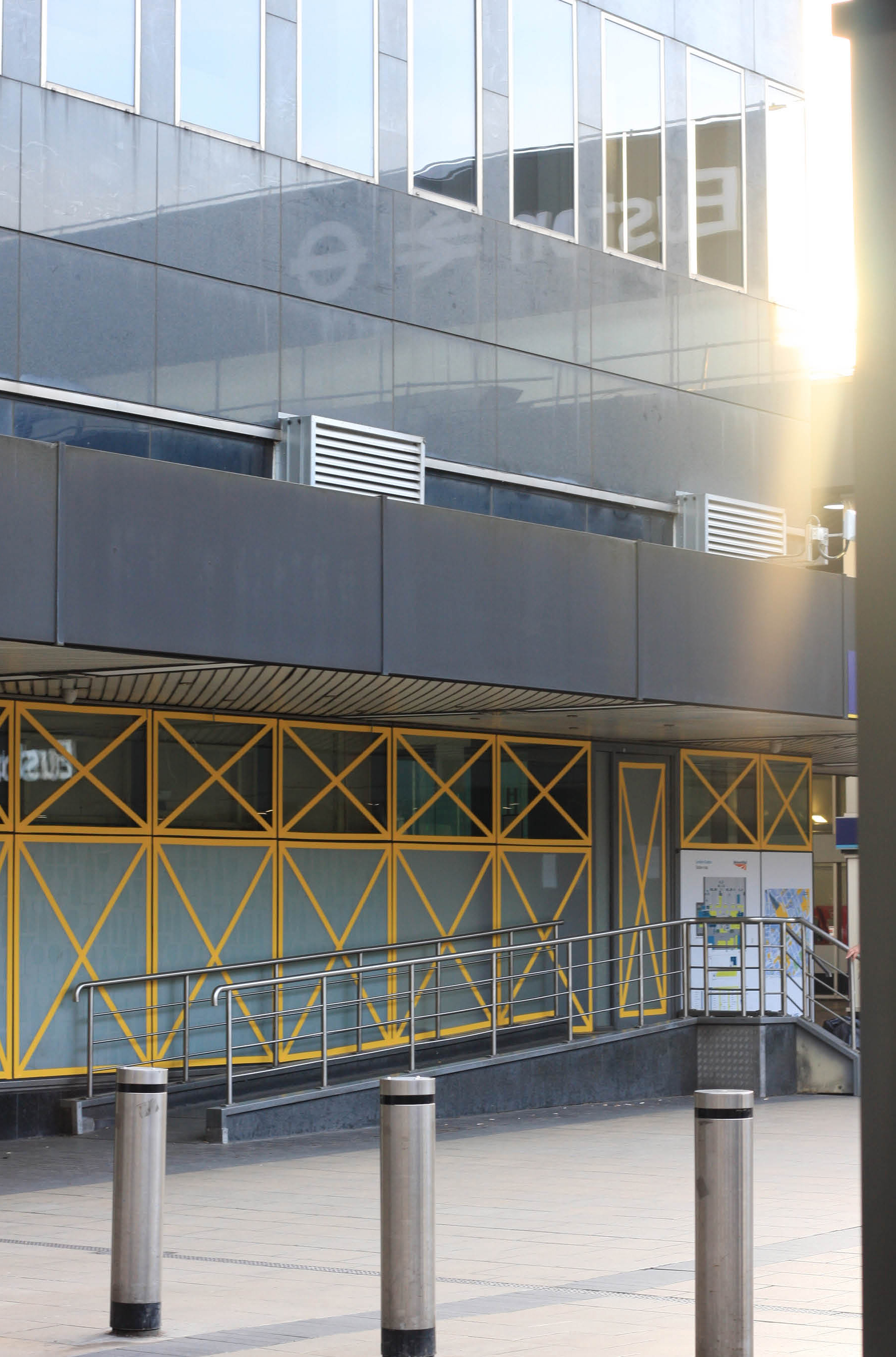 Port Portal Piscator' Architectural Public Art installation by  Henry Coleman of yellow vinyl crosses across the modernist façade of old Euston station forecourt building by Seifert and Partners, now destroyed, with blurred figures walking in the foreground 