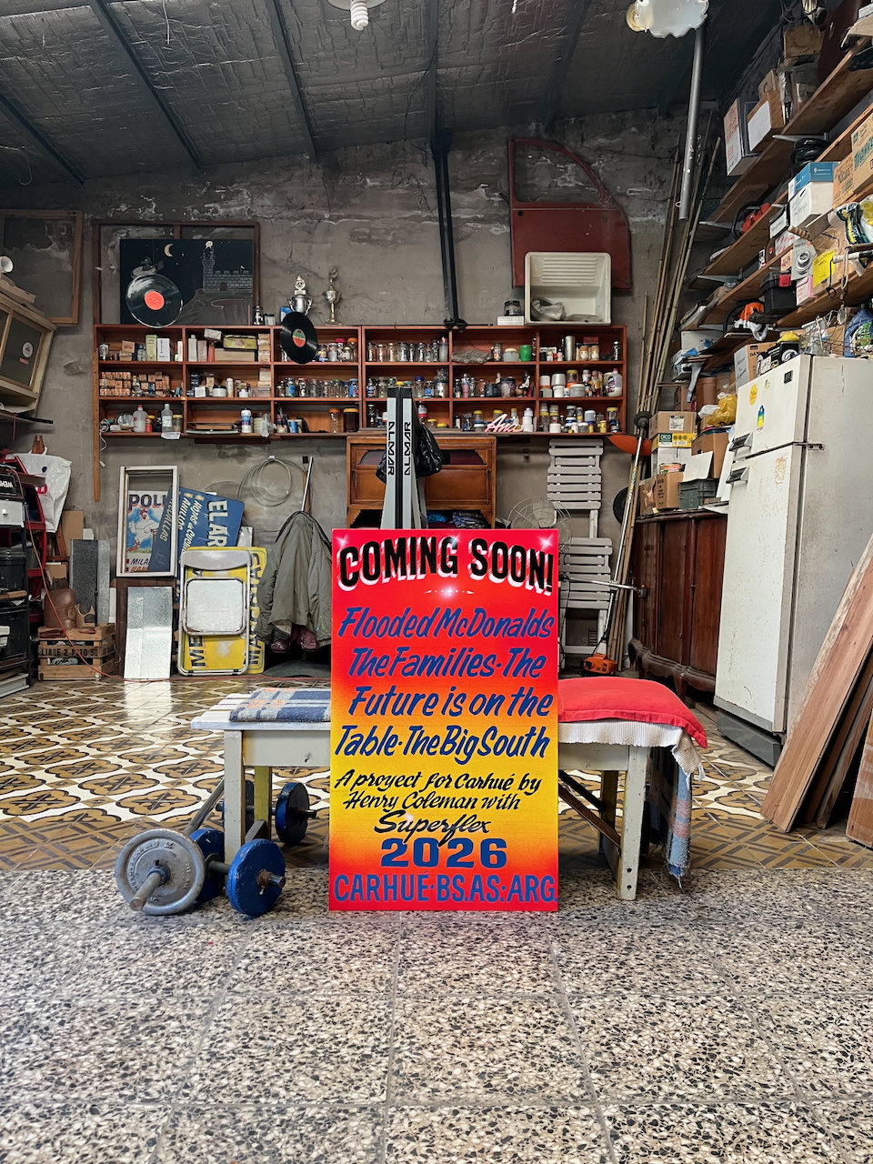 Coming Soon (2025) by Henry Coleman with Colo Maldonado: Red and orange text board leaning on a rustic wooden bench inside a cluttered artist's workshop.