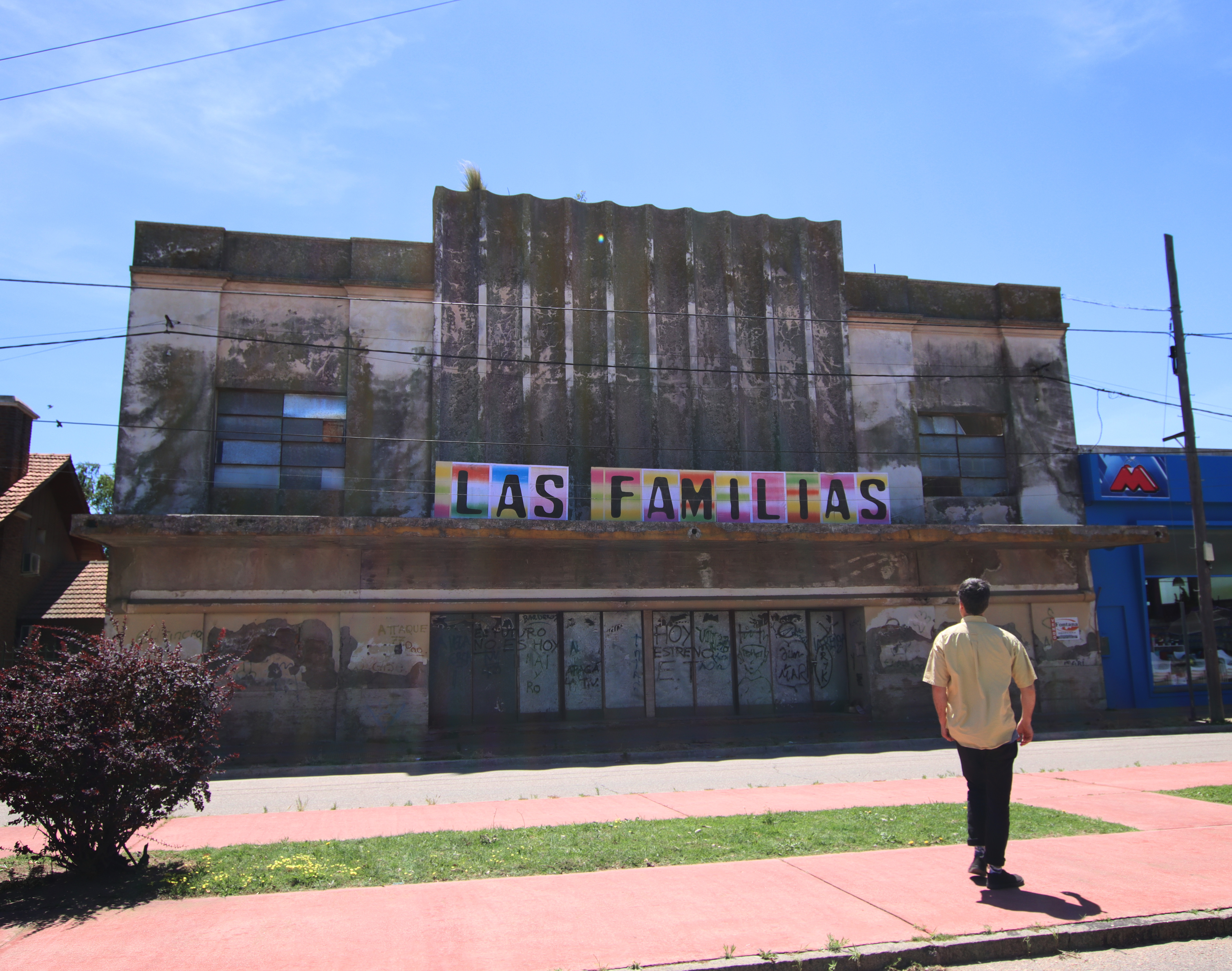 Las Familias (2025) by Henry Coleman: Man in yellow shirt walks towards the Poster Artwork installationgrand, distressed art deco facade of Cinema Grand Sud in sunlight. 