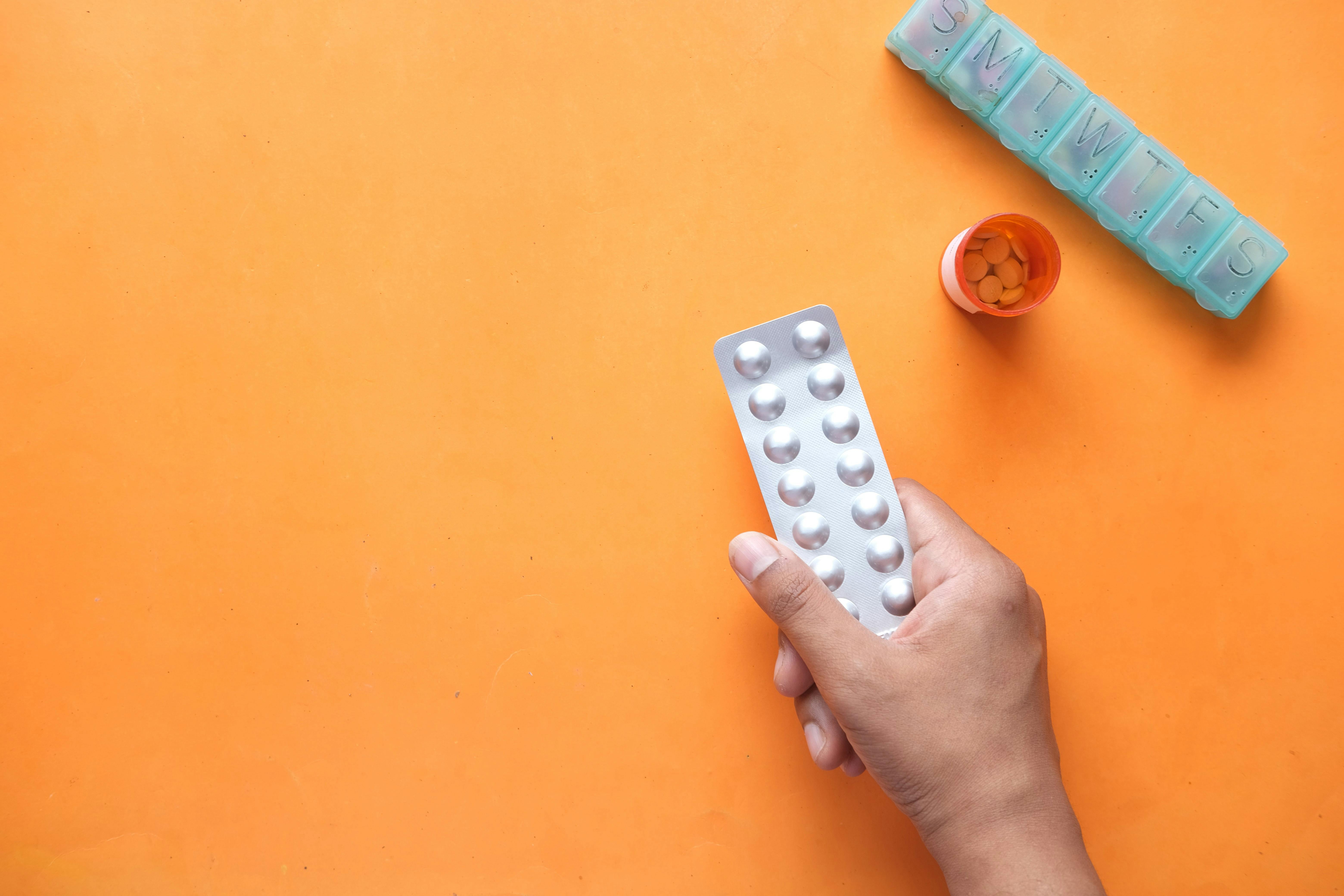 hand holding a blister black, with a filled pill box and pill background beside it on an orange background