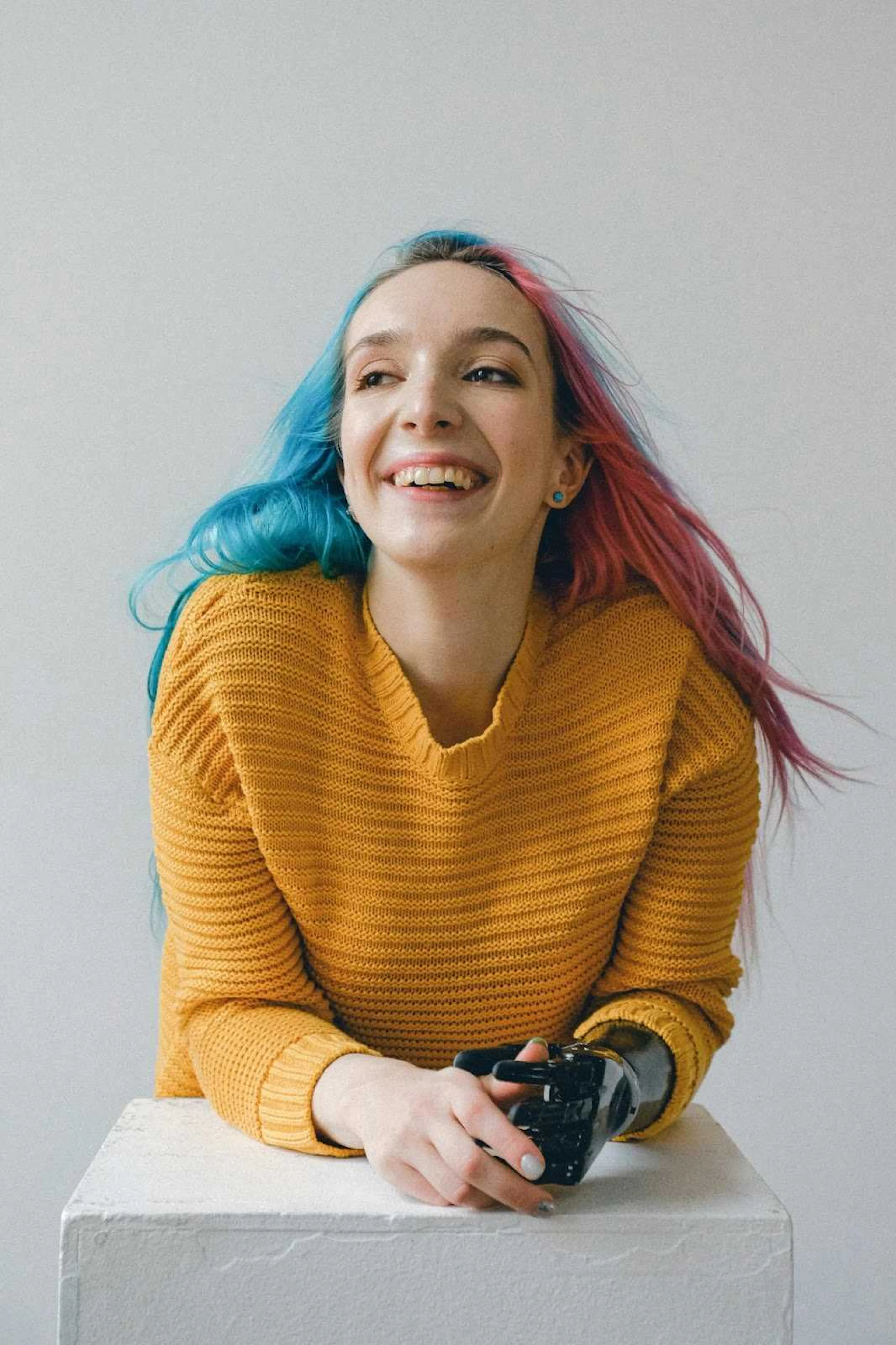 A woman with pink and blue hair, a yellow sweater, and a prosthetic arm poses and smiles while leaning on a white block