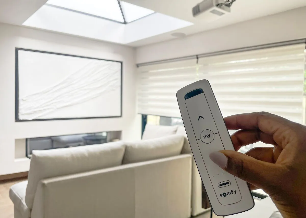 A hand holding a Somfy remote control to operate motorised pleated blinds in a skylit living room.