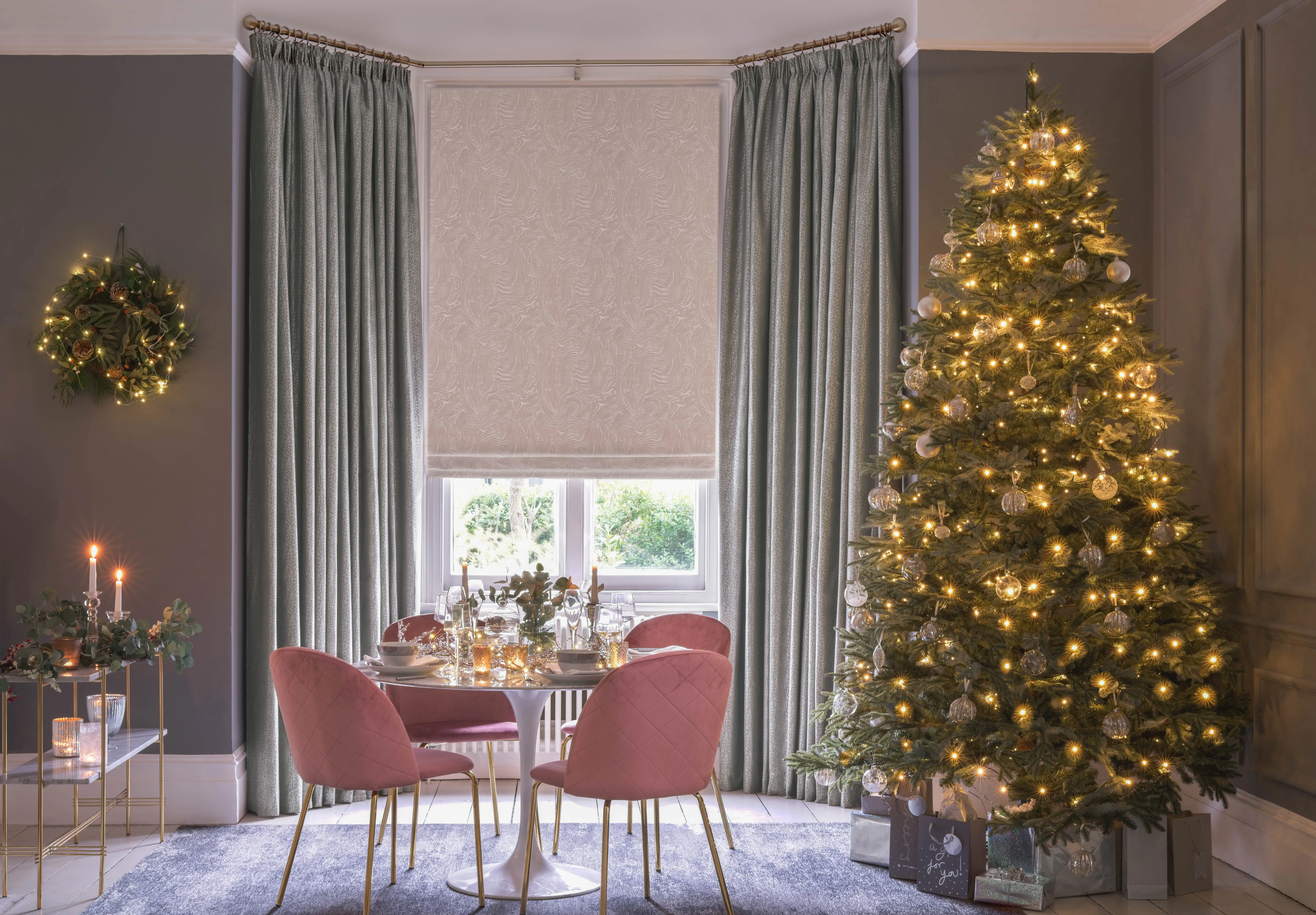 Bay window dressed with soft grey curtains and a Roman blind, overlooking a festive dining area and illuminated Christmas tree.