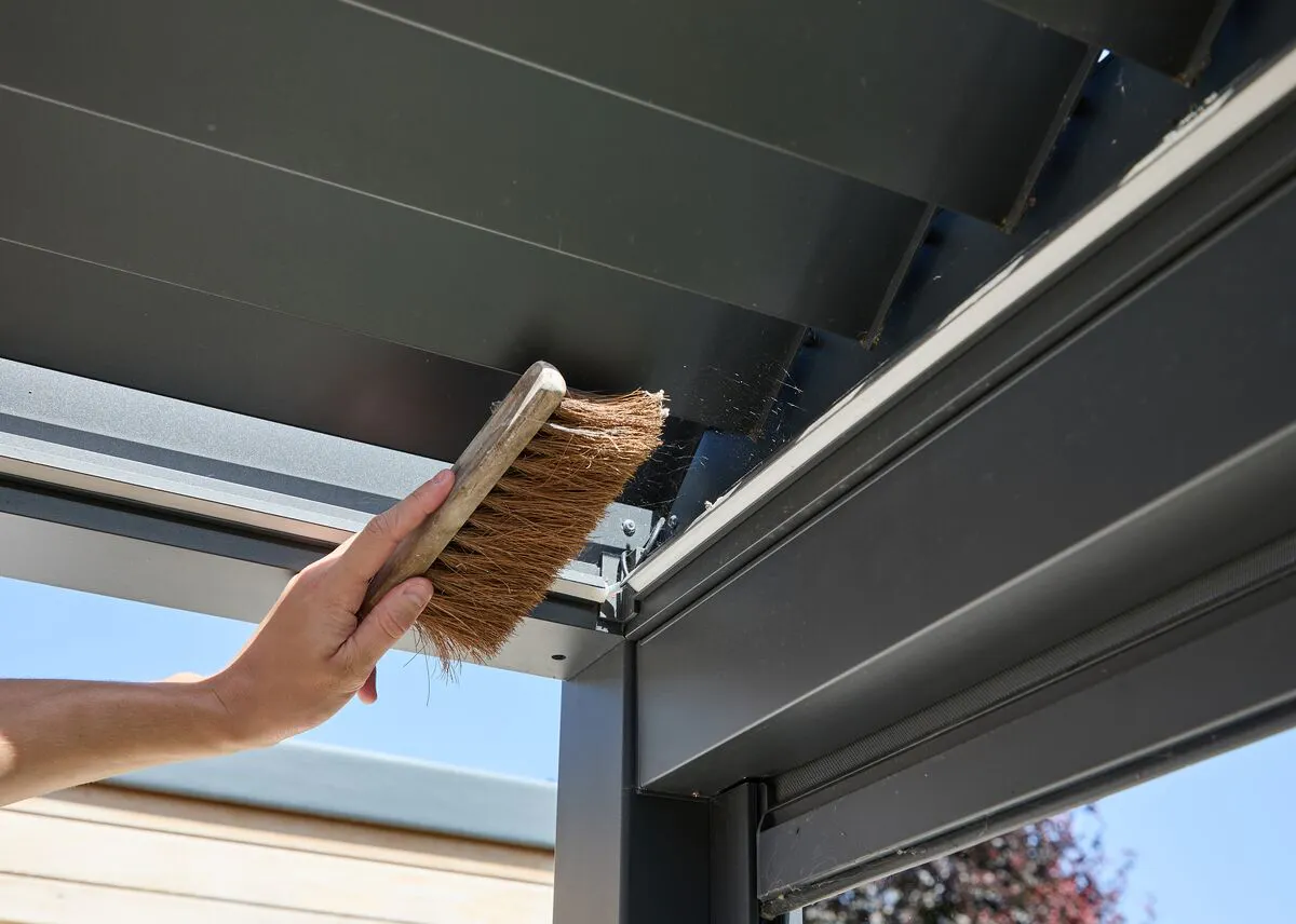 Person using a handheld brush to clean the louvred roof and frame of a modern aluminium pergola on a sunny day.