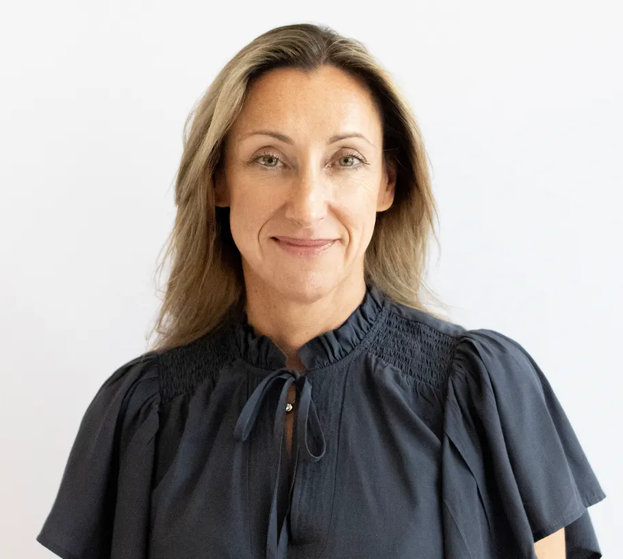 Studio headshot of a person with shoulder-length hair wearing a navy blouse against a light grey background.