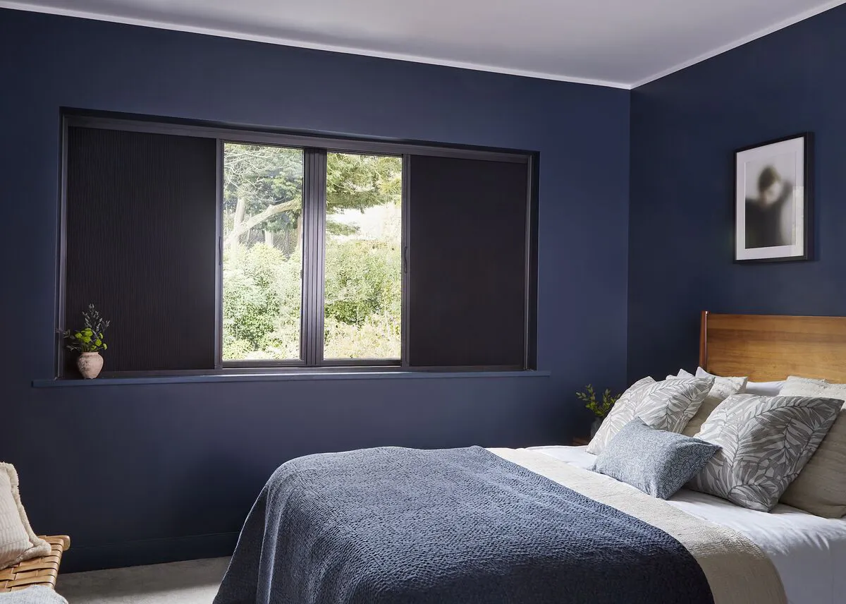 A navy blue bedroom with a white ceiling and black blackout screen blinds. Throws and cushions are layered on a bed with a wooden headboard.