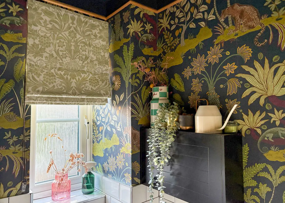 Olive-toned Roman blind at a frosted window, set against bold botanical wallpaper with plants and decorative accessories.