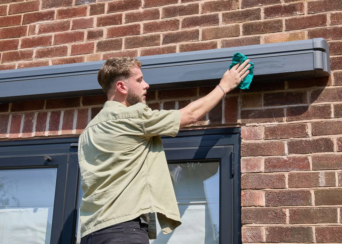 Person wiping down the cassette of a grey patio awning mounted on a brick house during routine outdoor maintenance.
