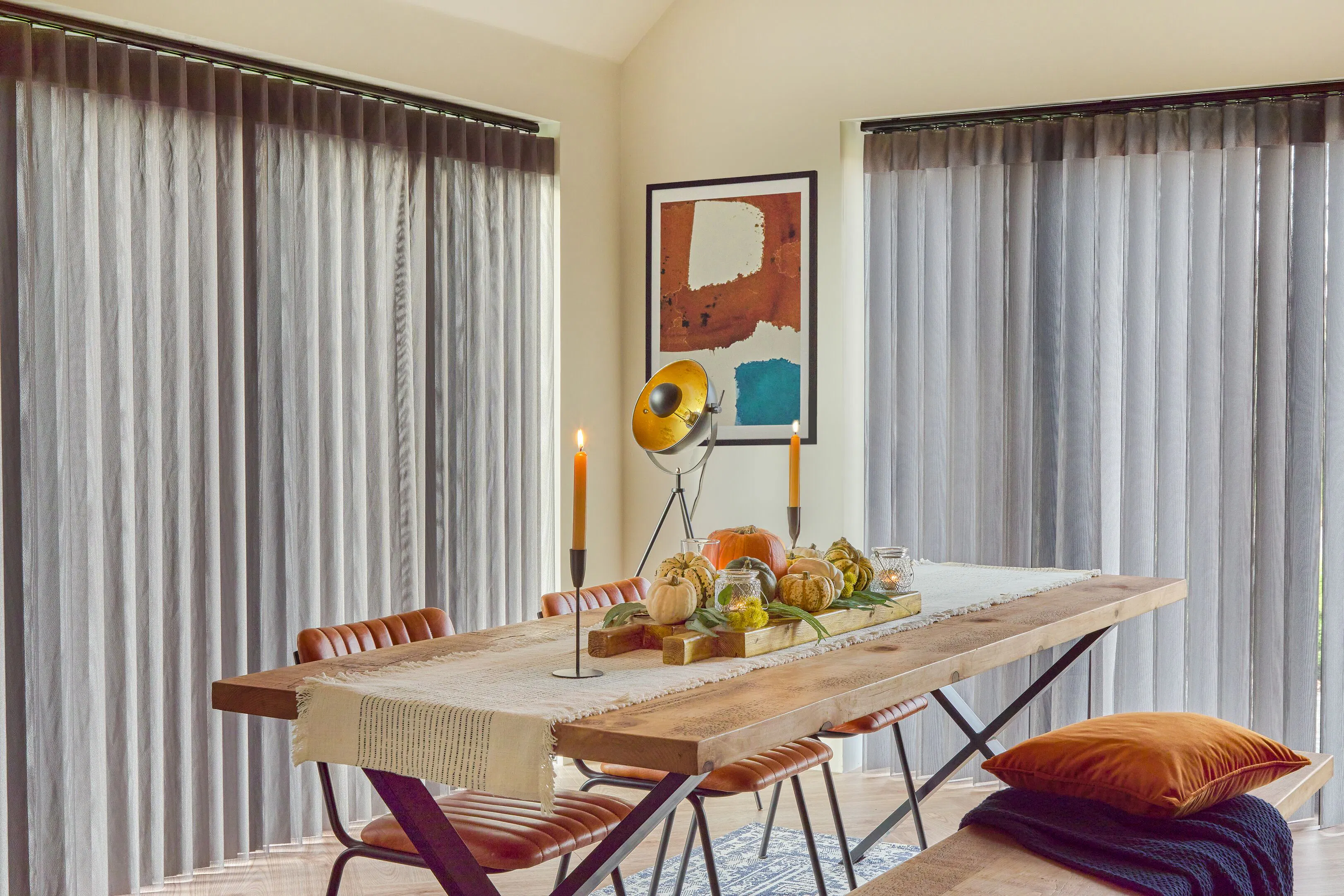 Grey Diffused blinds frame two large dining room windows, filtering daylight beside a rustic table styled with autumnal decorations.