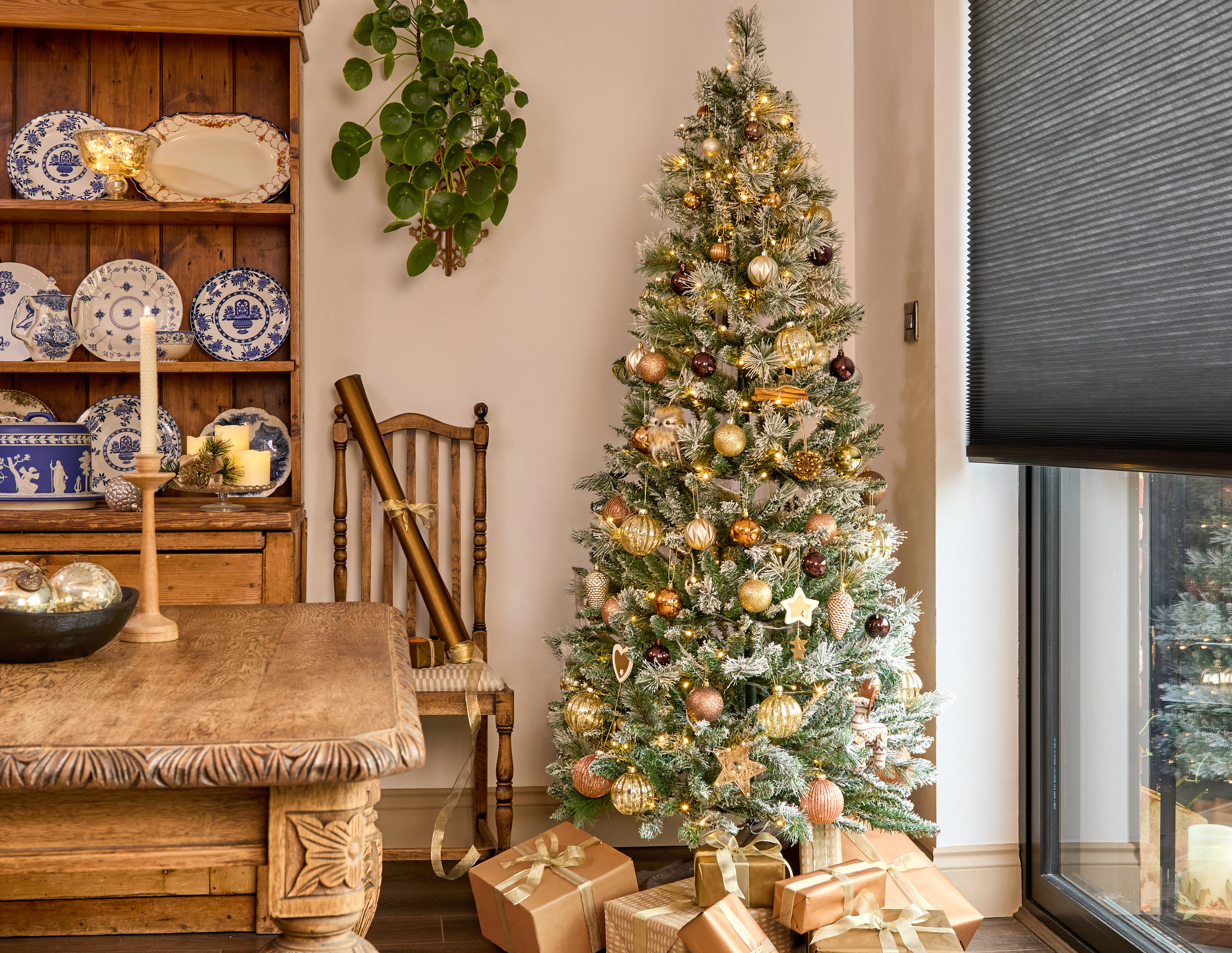 Frosted Christmas tree beside a large window fitted with a dark Pleated blind, surrounded by wrapped gifts in a rustic dining room.