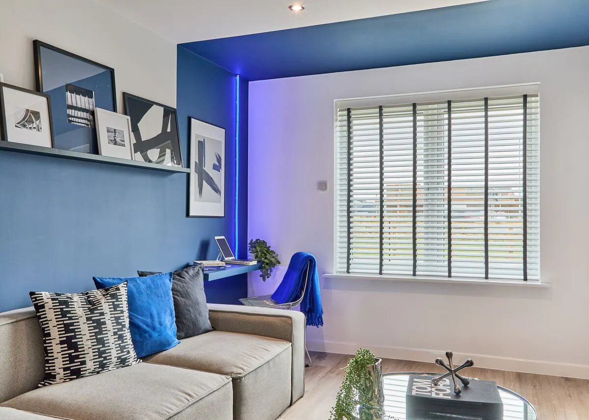 A blue living room with a Wooden blind, black-and-white artwork and a neon blue accent light. A small desk is tucked into the corner next to a light-coloured sofa.