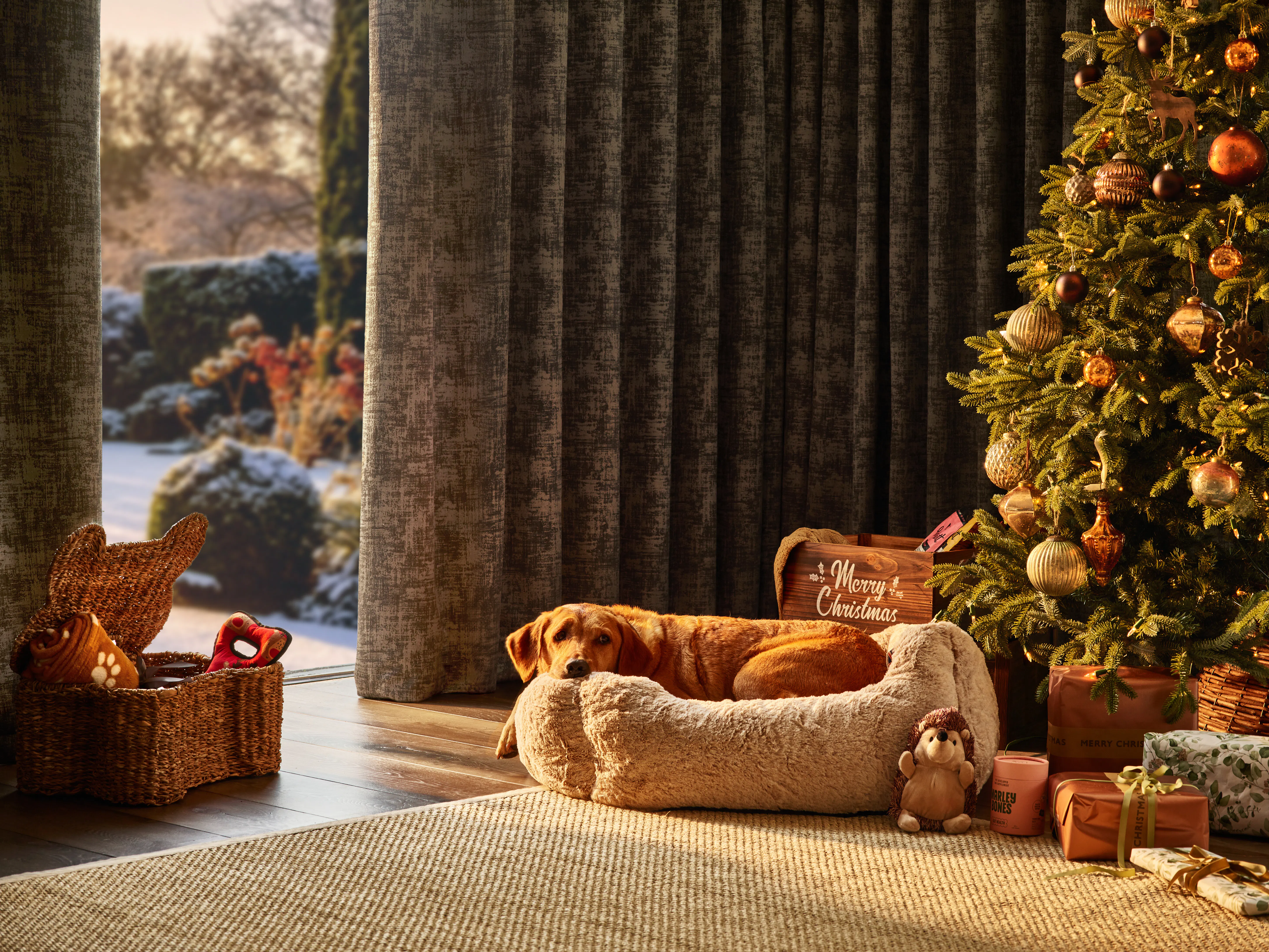 Floor-to-ceiling dark curtains frame a snowy garden view beside a decorated Christmas tree and cosy dog bed.