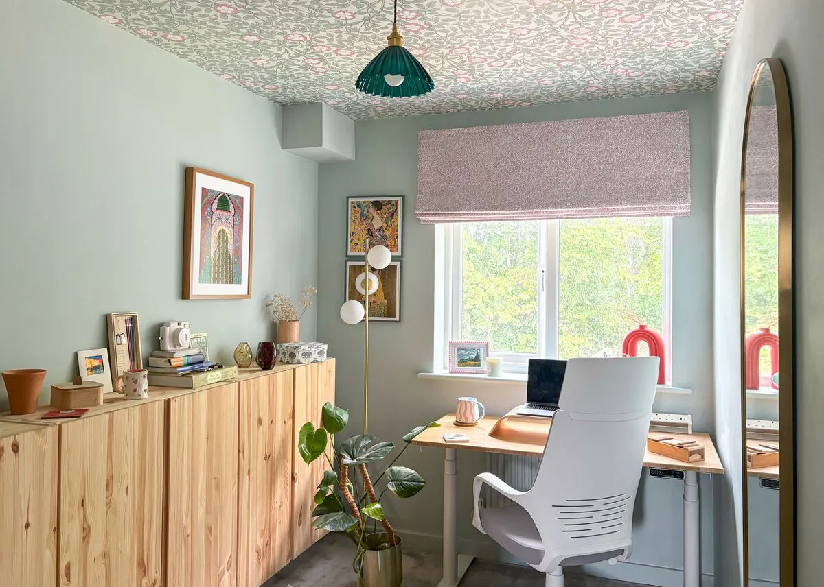 Sage-green home office with floral wallpapered ceiling, dusky pink patterned Roman blind and pine storage units.
