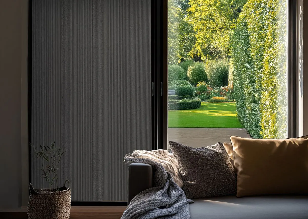 A sofa in front of wide patio doors, half covered by a blackout blind. The room is in shadow, but a sunny landscaped garden can be seen through one door.