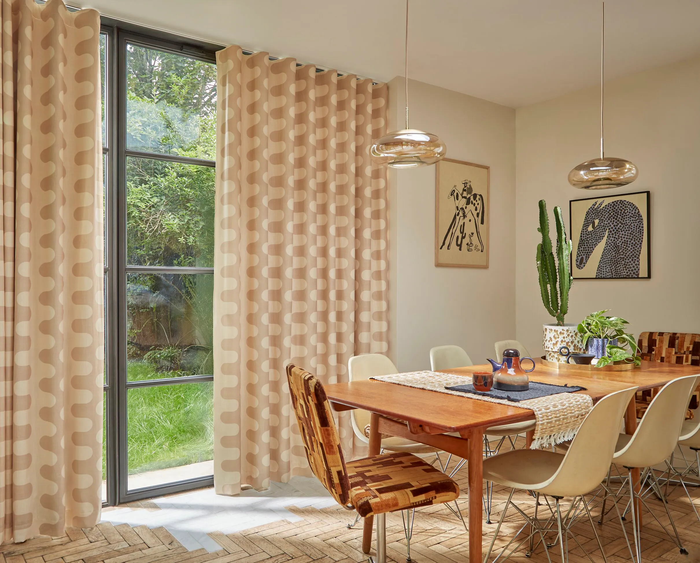Patterned blush curtains frame a tall black-framed window, bringing soft light into a stylish dining area with modern furnishings.