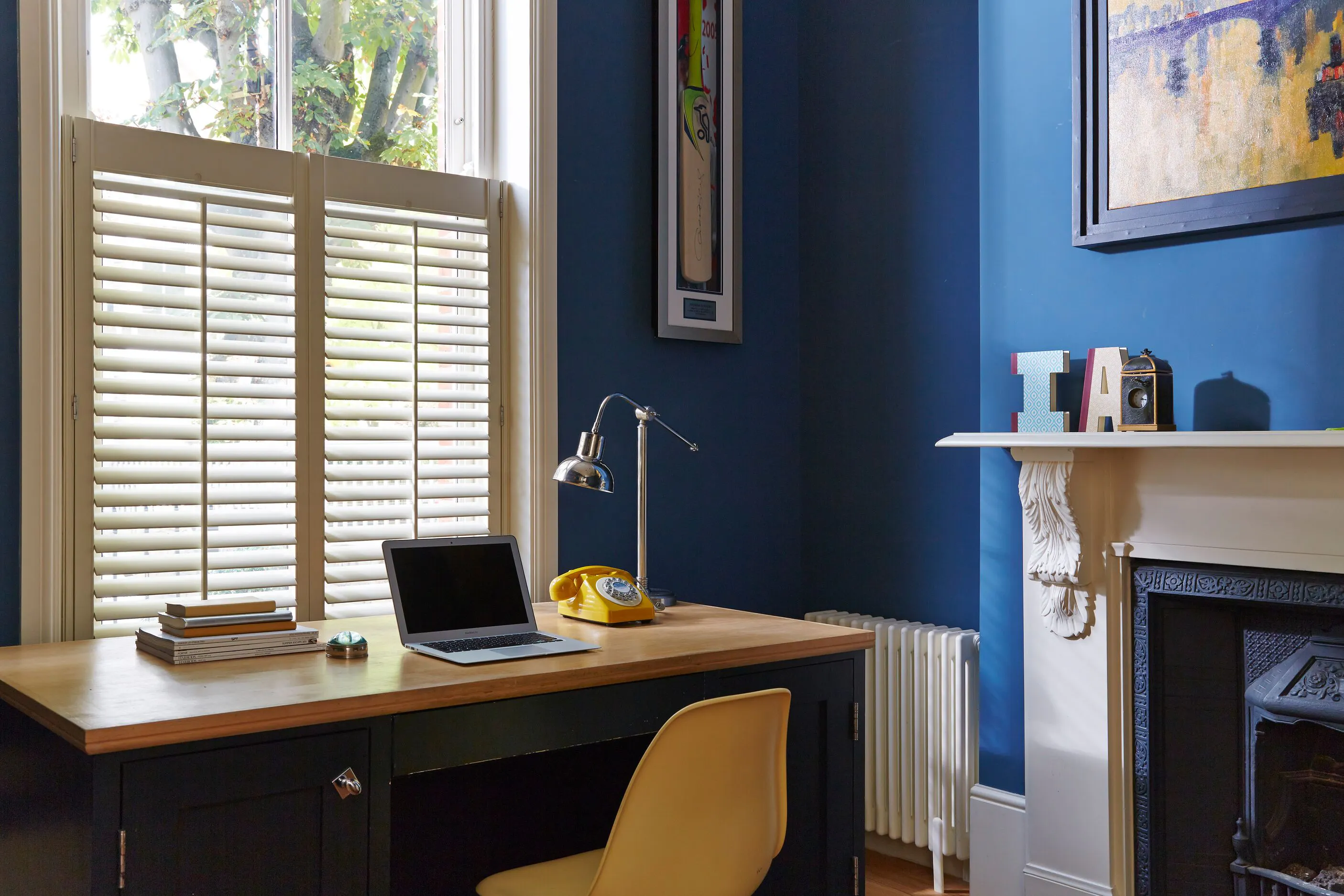 Home office with white window shutters, a wooden desk and laptop, set against deep blue walls beside a decorative fireplace.