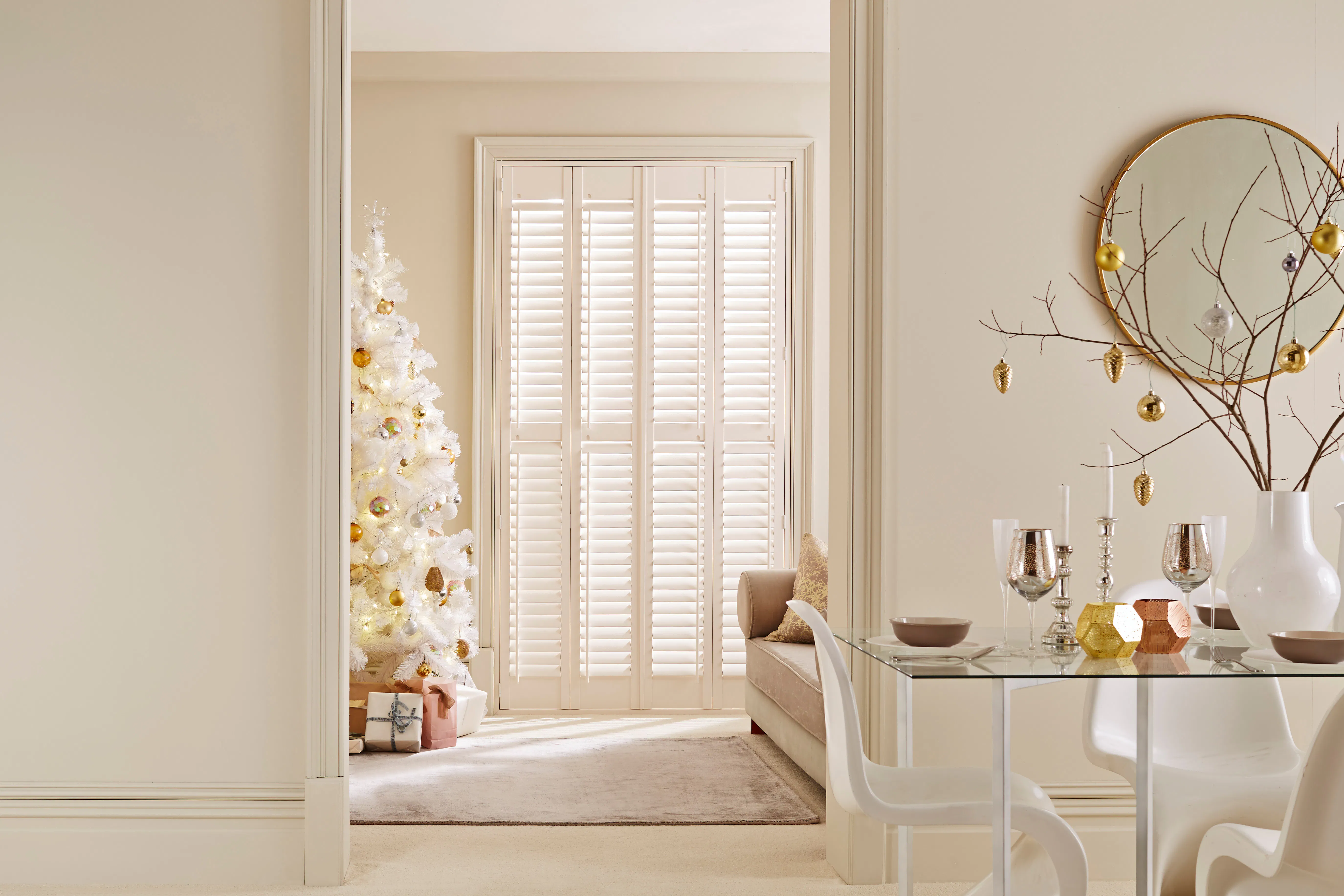 A neutral, minimal dining room with a glass table styled with a vase of bare twigs hung with delicate gold ornaments. An open doorway leads to a second room, featuring a white Christmas tree and glazed door behind cream-coloured shutters.