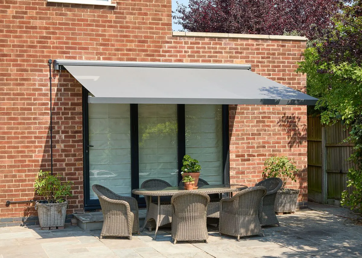 Contemporary patio awning extended over an outdoor dining set beside a brick house, creating shade across the seating area.