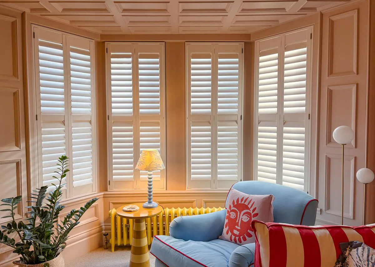 Bay window fitted with white shutters, filtering soft daylight into a cosy seating area with a blue armchair and colourful cushions.