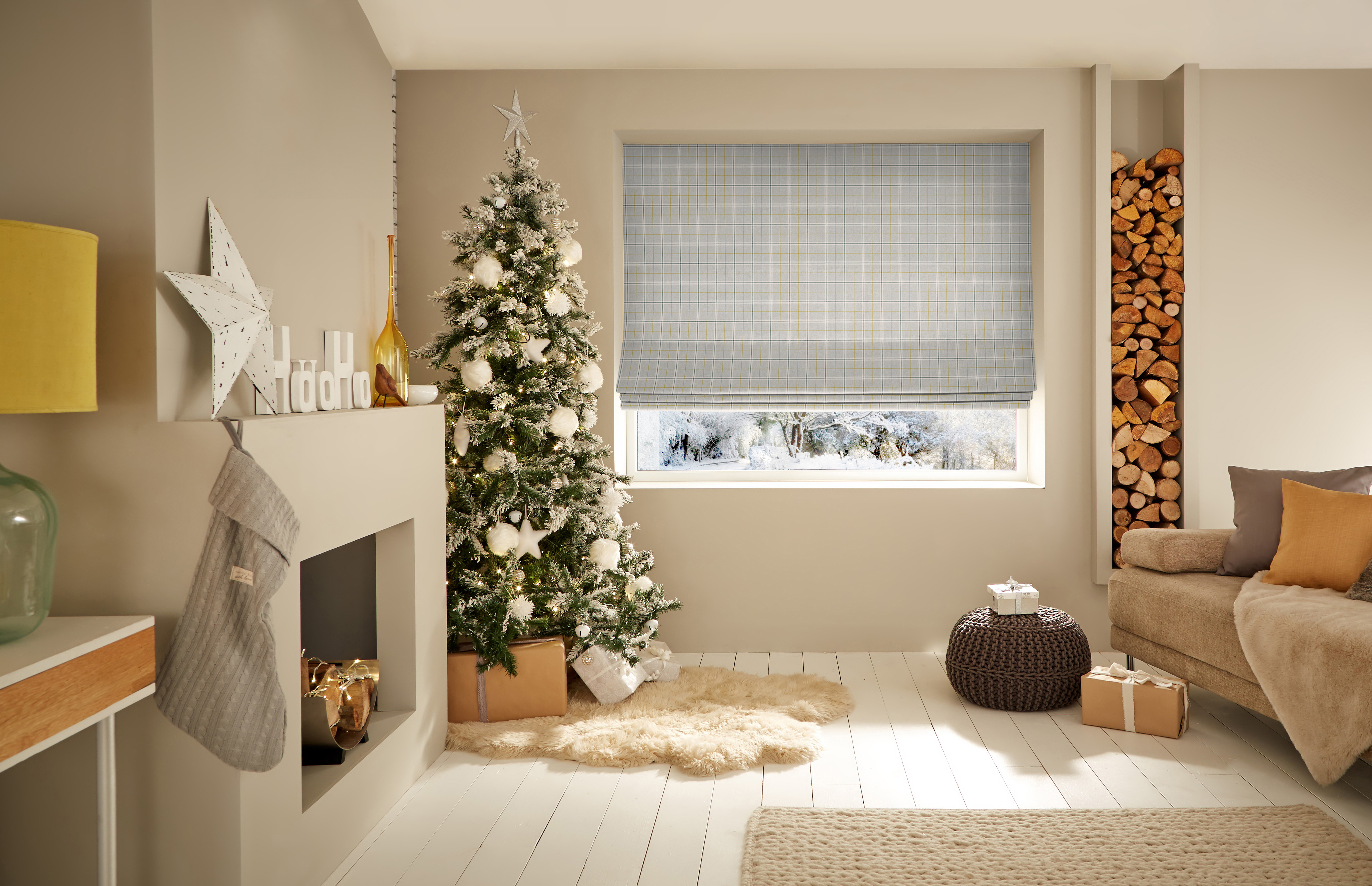 Bright living room with a wide window fitted with a checked Roman blind, beside a snow-dusted Christmas tree and fireplace.
