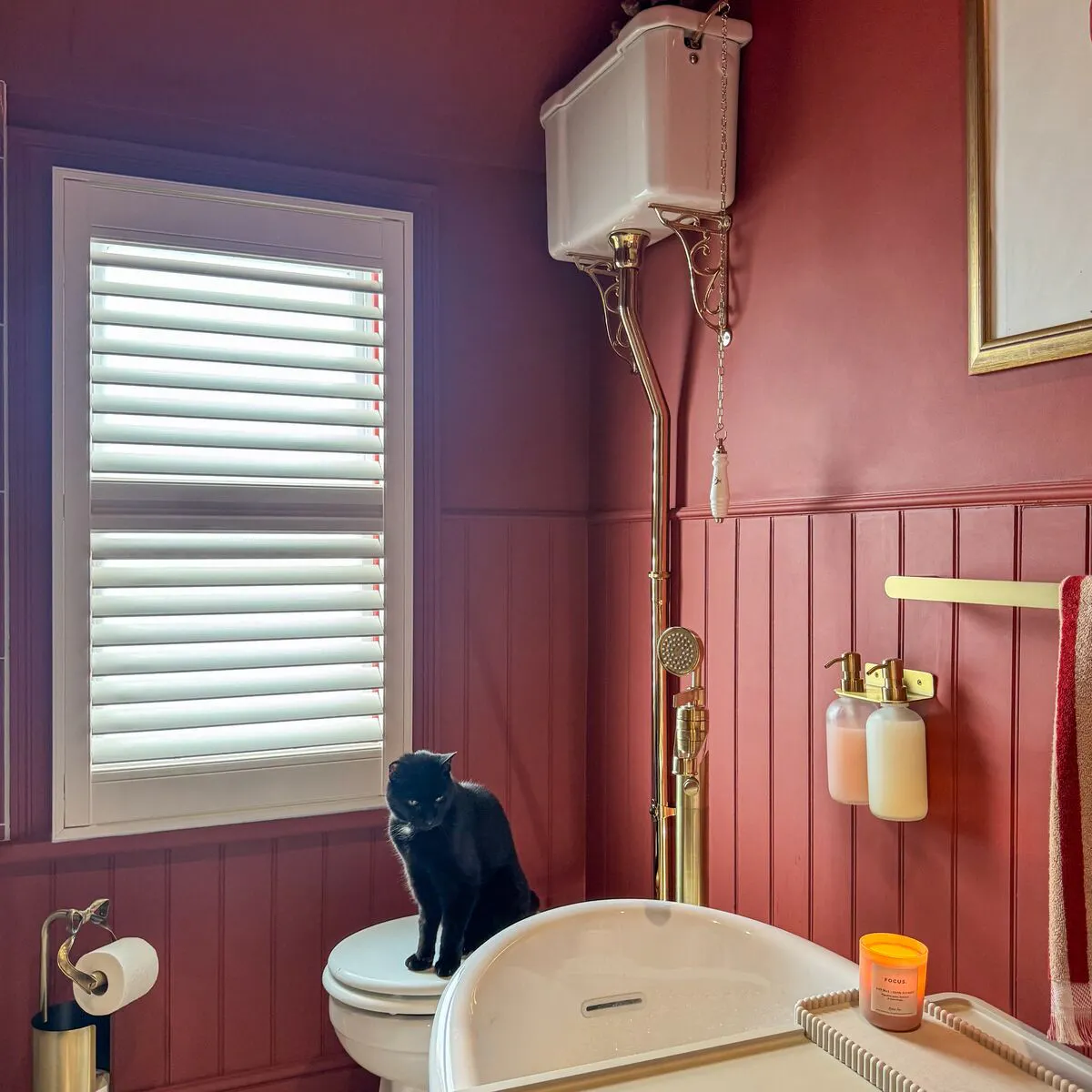 White shutter at a bathroom window, paired with rich red wall panelling, a freestanding bath and brass fixtures.
