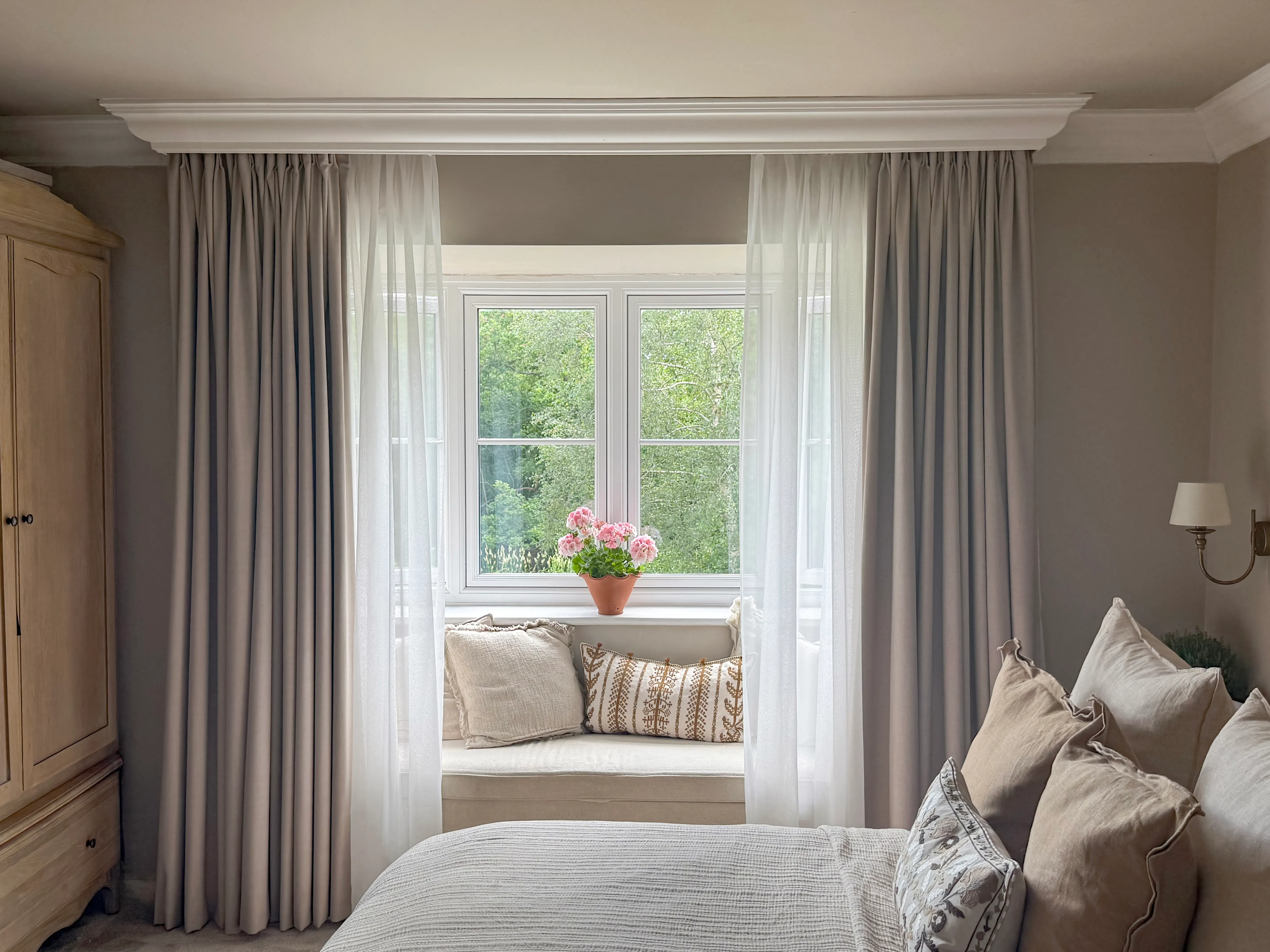 Bedroom window with sheer and floor-length curtains framing a cushioned window seat, overlooking leafy greenery outside.