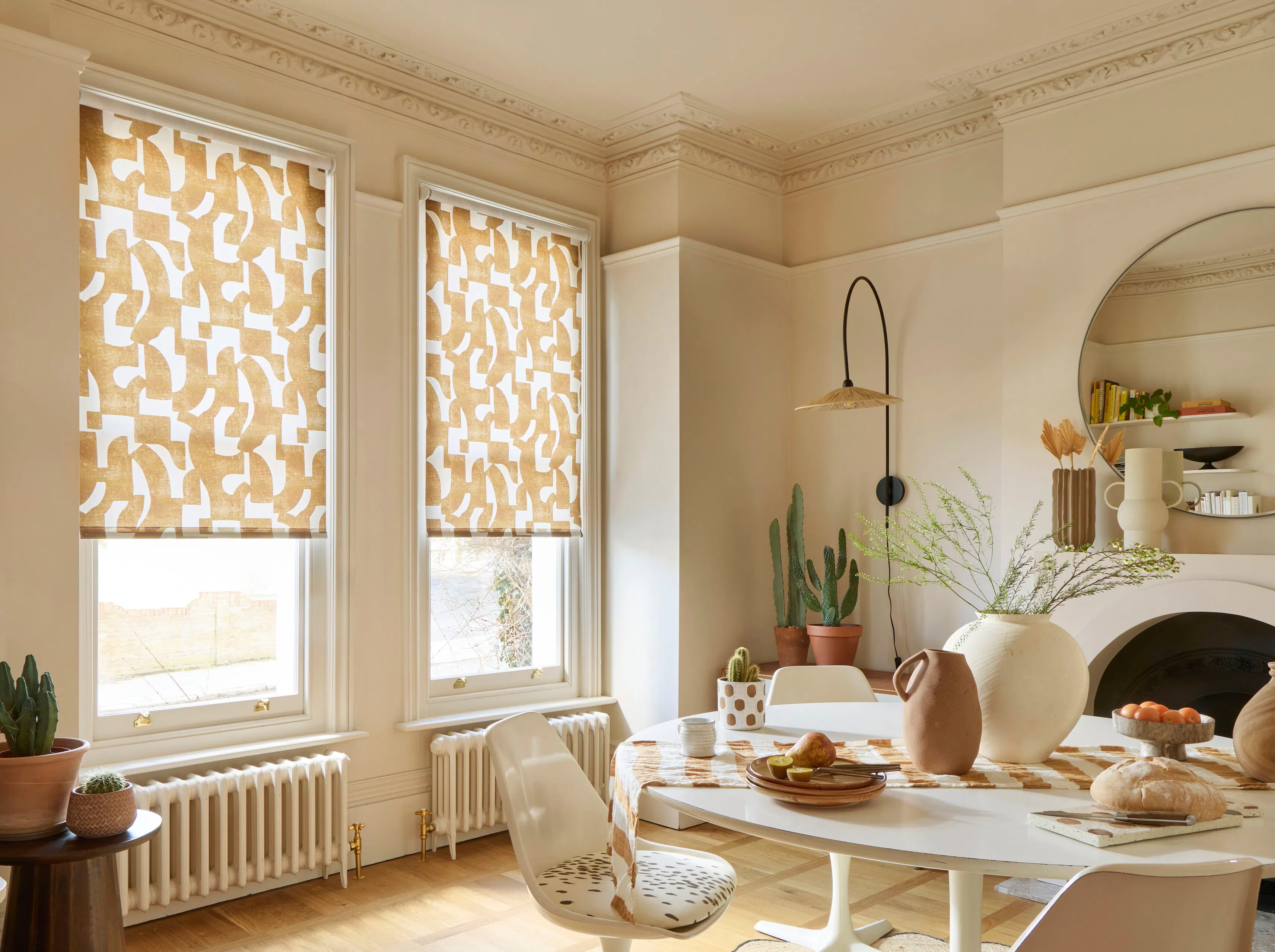 Two tall windows with patterned Roman blinds filtering daylight into a bright dining room with ornate cornicing, plants and a round table.