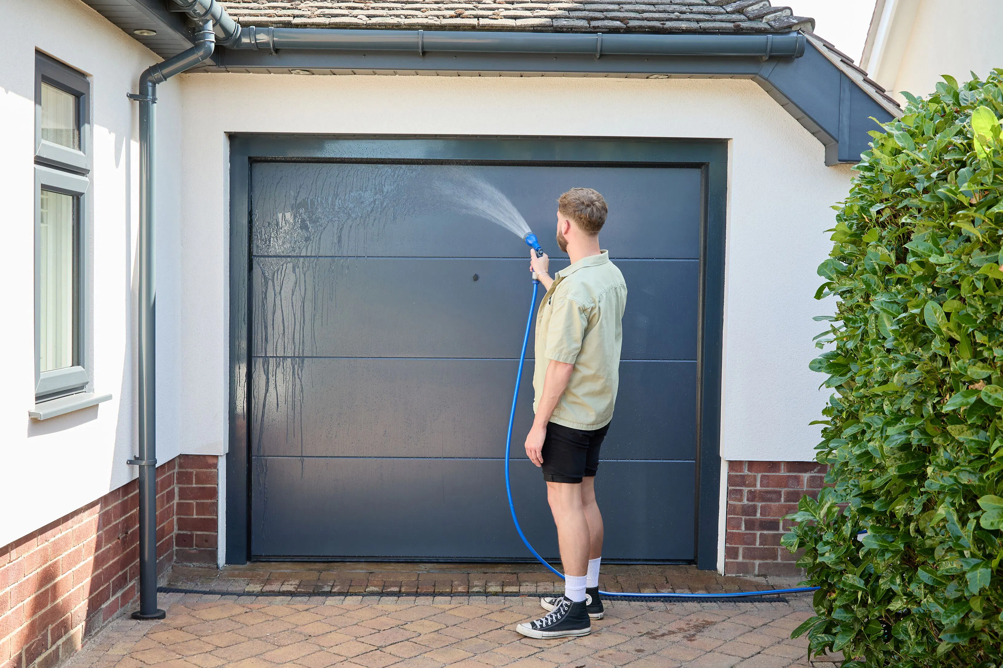 Person rinsing an anthracite grey garage door with a hose outside a house, with front window, white walls and paved driveway.