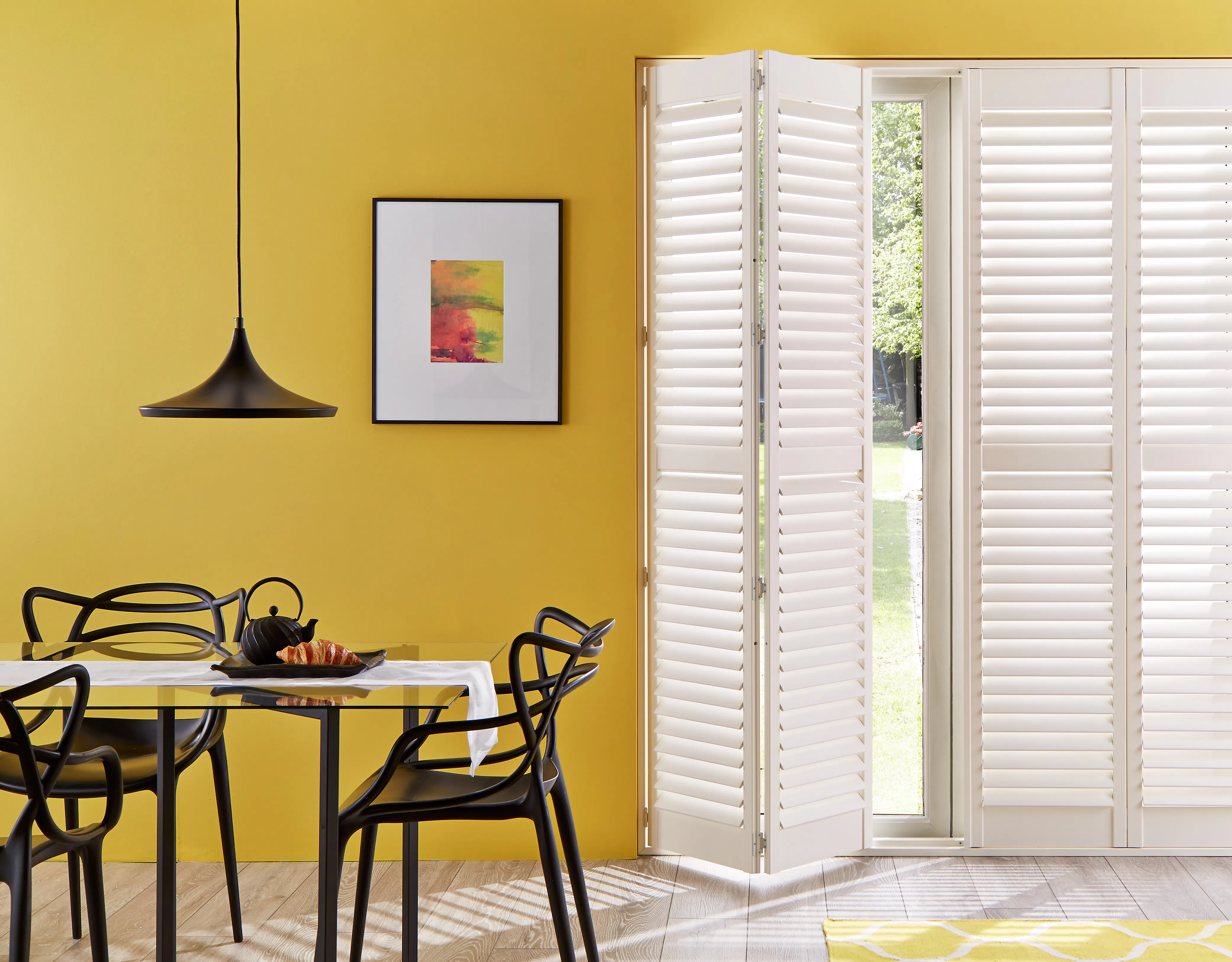 A bold, sunlit dining room with mustard yellow walls, white full-length shutters on patio doors and sculptural black dining chairs.