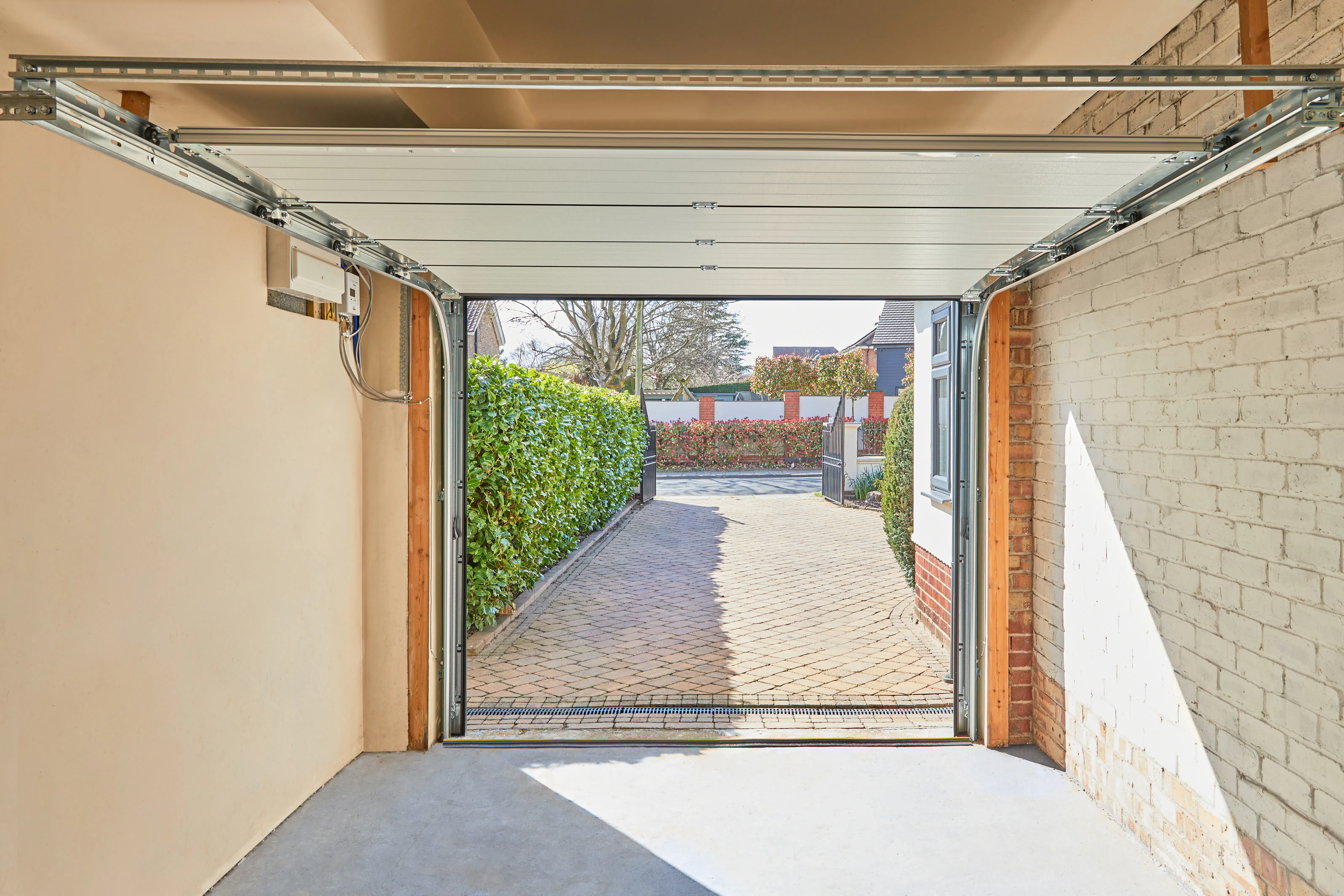 The view from inside a garage looking out through an open sectional door onto a block-paved driveway, hedging and the street beyond.