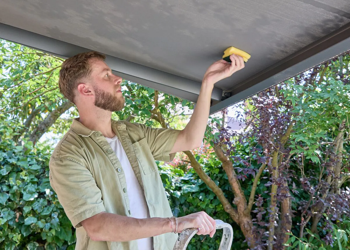 Person using a sponge to clean the underside of an awning while standing on a stepladder in a leafy garden.